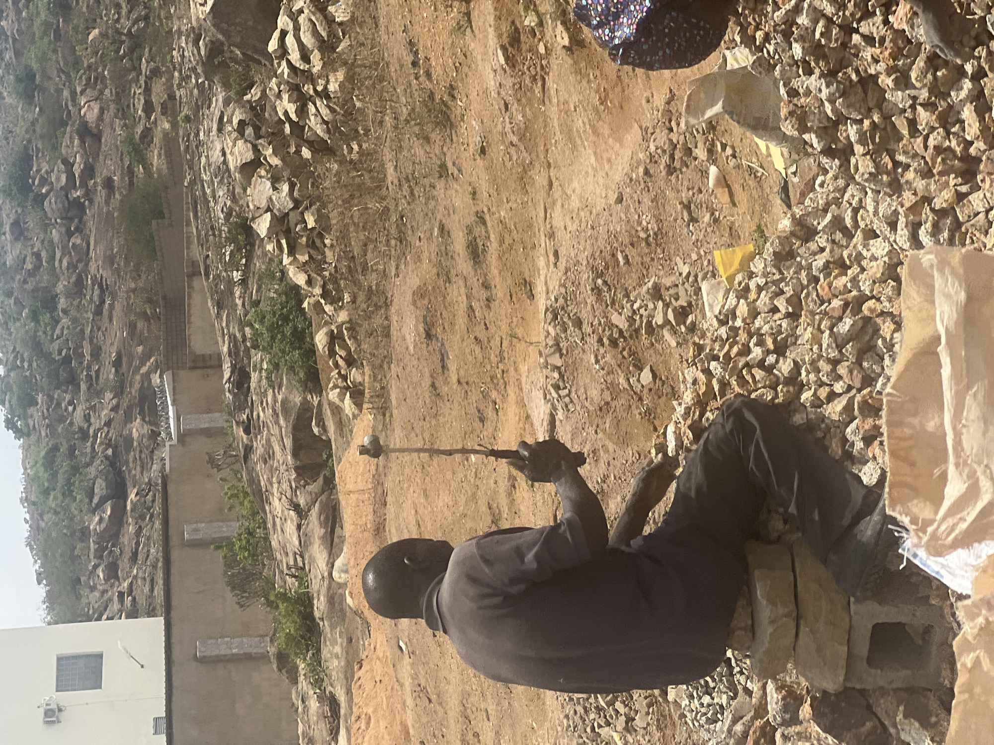 The image depicts an outdoor scene under bright daylight in a rocky, arid environment, identified as Gura Top, Nigeria. The sun is high, casting distinct shadows.
In the foreground, a dark-skinned man with a shaved head is seated on two concrete blocks, facing away from the viewer. He wears a dark short-sleeved top and dark trousers. He is holding a long wooden stick with a rounded, light-colored head, appearing to be a tool, possibly for breaking or digging. The ground immediately around him is covered in loose, light-colored gravel and dry, reddish-brown earth. A large, light-colored sack is partially visible next to his right leg. A portion of another person, showing a leg clad in colorful, patterned fabric, is visible in the upper right part of the frame.
The mid-ground and background are characterized by rugged, rocky terrain featuring numerous large rock formations and outcroppings. Sparse green vegetation, including small bushes and shrubs, is scattered across the landscape. Several structures are visible, integrated into the rocky surroundings, including a white building with a dark-framed window and an air conditioning unit, along with other concrete or stone constructions, suggesting a settlement or ongoing construction activity. The overall impression is a dry, sun-drenched landscape with signs of human activity. No discernible text is visible in the image.