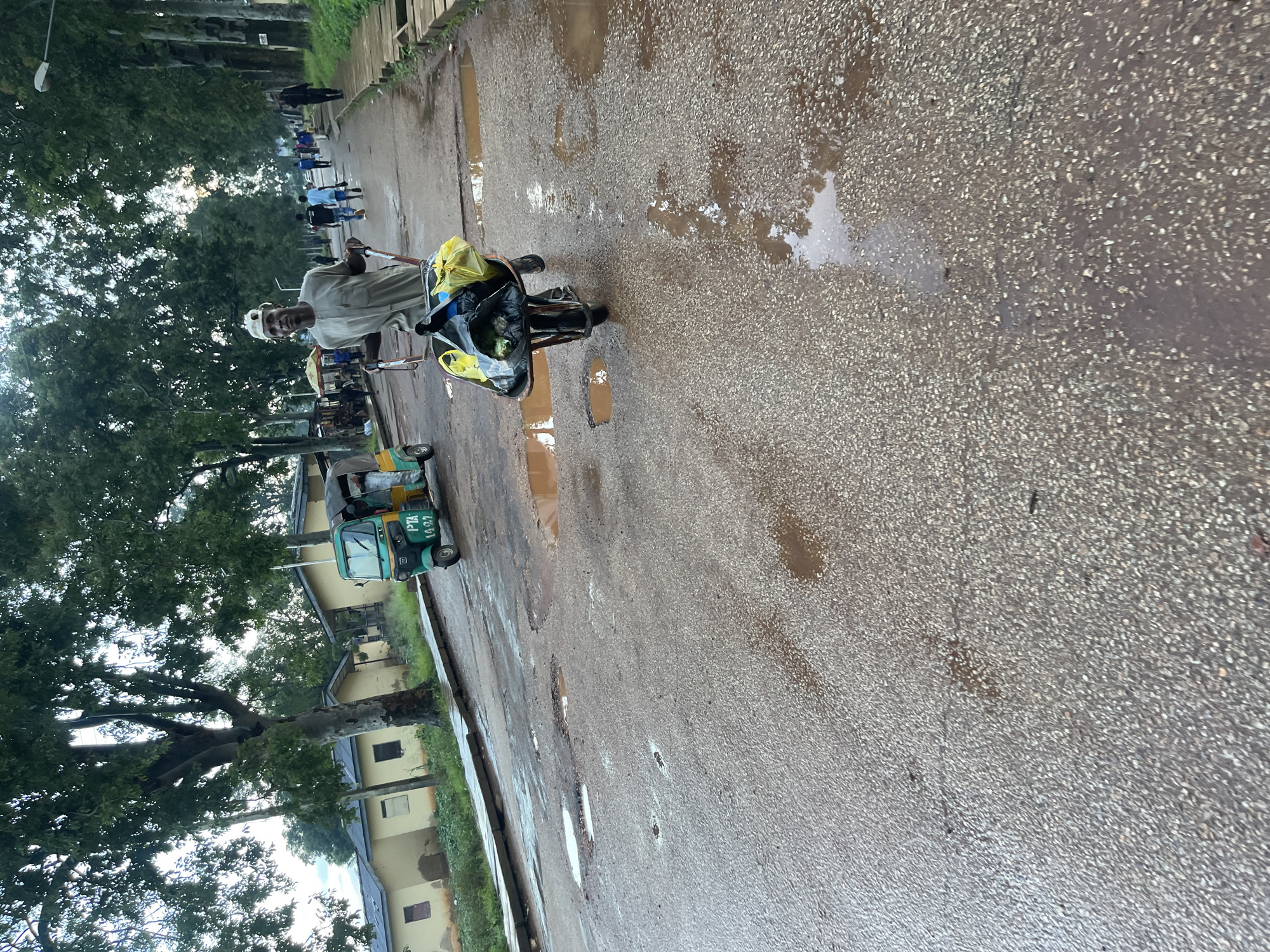A man wearing a light-colored tunic and cap is pushing a wheelbarrow loaded with bags on a wet asphalt road. The road is lined with trees on the right and buildings on the left. In the background, a three-wheeled vehicle, possibly a tuk-tuk, is parked on the side of the road, and other people are walking along the road and sidewalk. Puddles of water are visible on the road surface, indicating recent rain. The scene appears to be an outdoor environment in a populated area. The location is identified as Rafin Par, Nigeria.