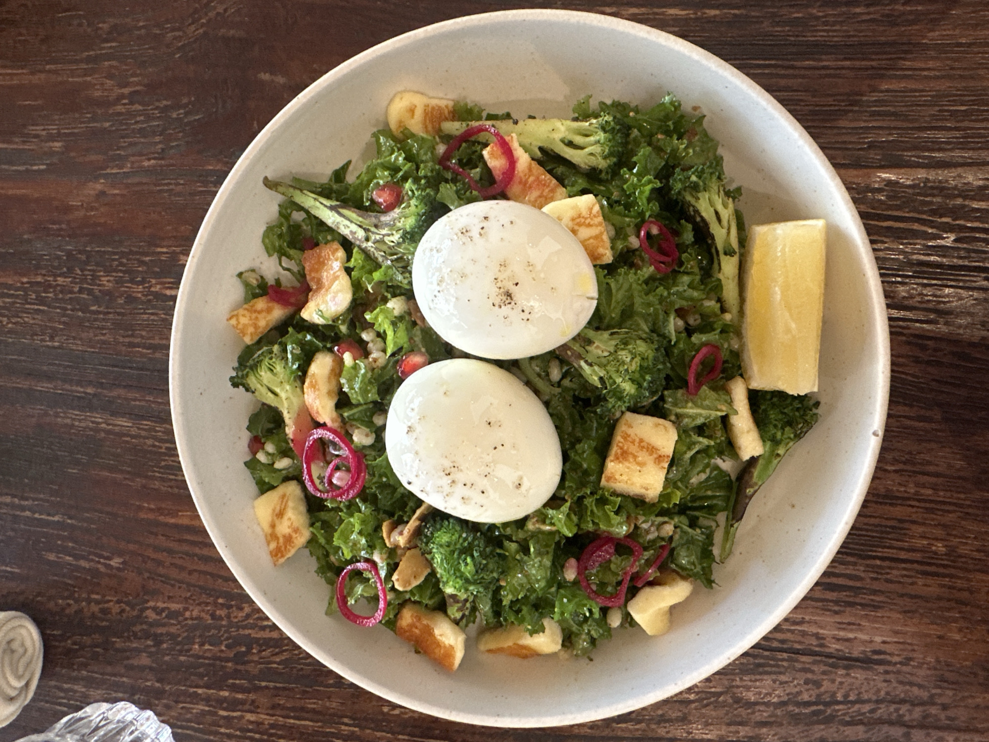 A top-down, eye-level shot captures a vibrant salad served in a light-colored, circular bowl set against a dark, wooden table. The salad is a medley of ingredients, featuring chopped green kale as the base, interspersed with florets of broccoli, cubed pieces of grilled halloumi cheese, pomegranate seeds, and thinly sliced red onion rings. Two halves of a hard-boiled egg, lightly seasoned with pepper, are centrally placed. A wedge of lemon rests on the side of the bowl. The lighting is natural and bright, highlighting the textures and colors of the food. The background is out of focus, but hints at a textured, possibly woven napkin and a glass to the upper left.