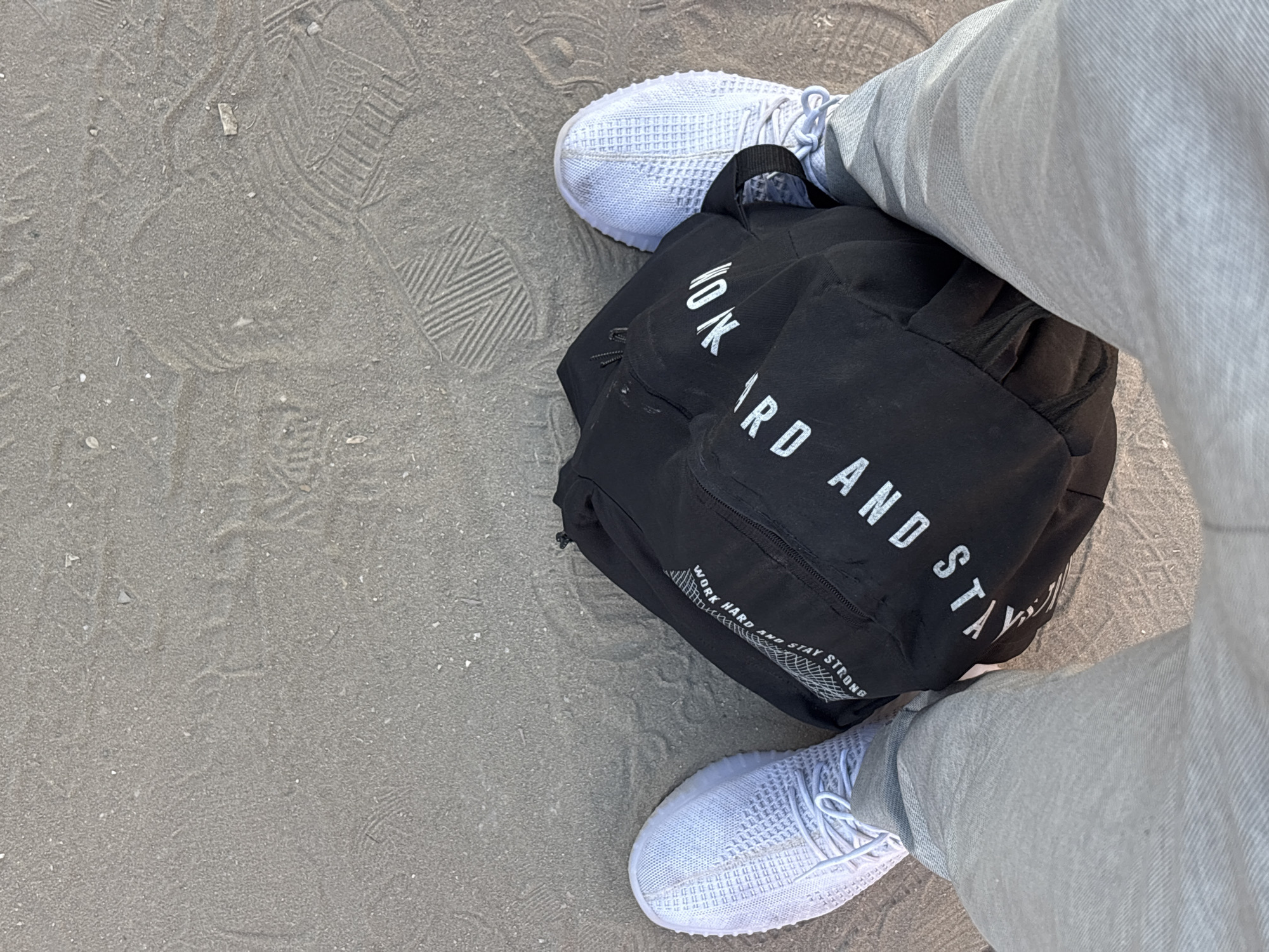 This media file shows a close-up, overhead view of a person's legs and feet, along with a black backpack, on a sandy surface. The person is wearing light grey pants and white athletic shoes. The backpack is resting on the sand, and appears to be made of a fabric material. White text on the backpack reads "WORK HARD AND STAY STRONG". The sandy surface shows imprints of what appear to be shoe prints. The lighting suggests it is daytime, and the overall scene appears casual and outdoors. The location is identified as Gaza, Palestinian Territories.