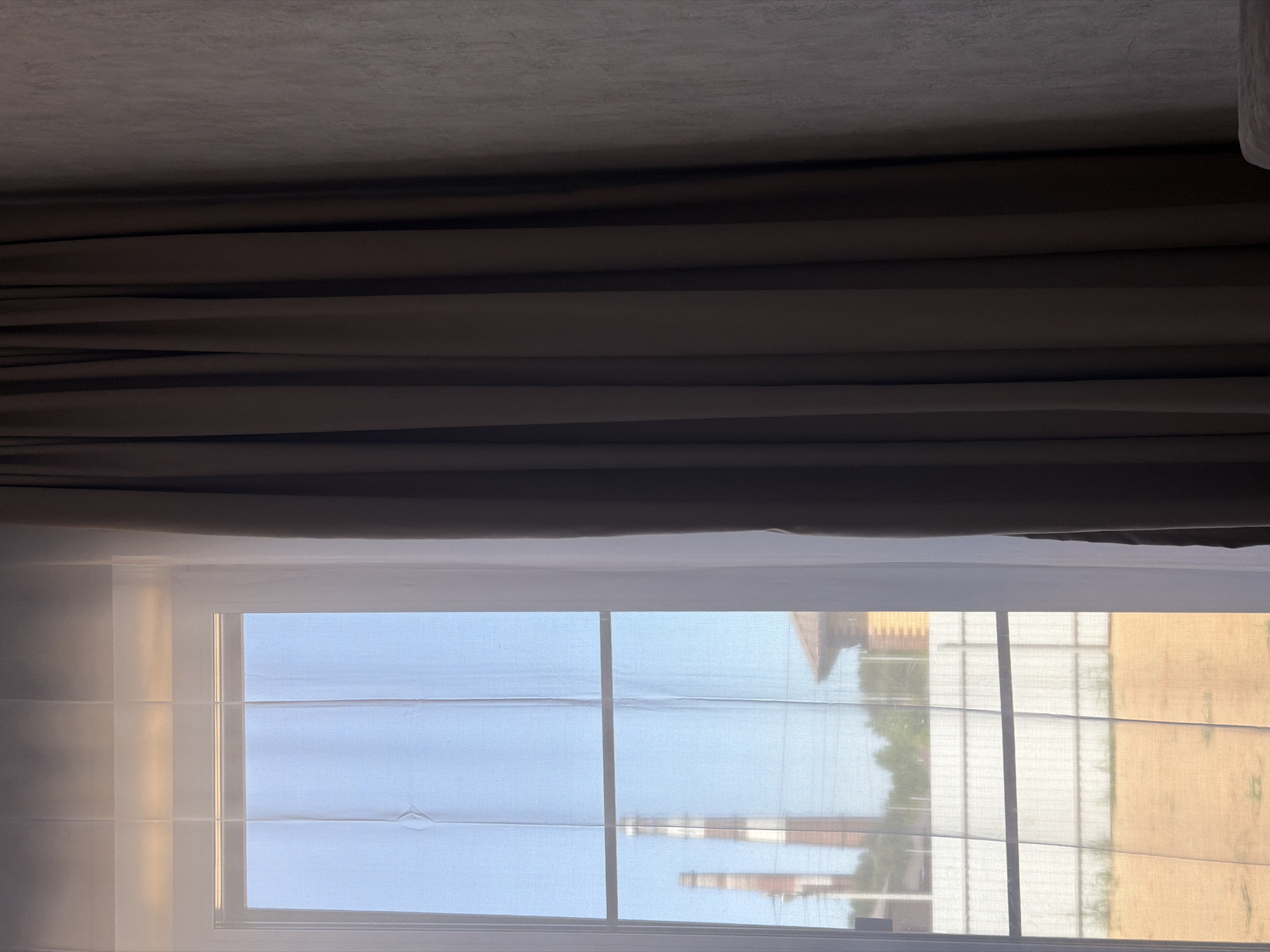 The image shows a view from inside a room, looking out through a window covered with a translucent blind. A heavy, brown curtain is draped above the window, partially obscuring the ceiling. The window offers a view of the outside, where one can discern elements of a cityscape. There is vegetation, a fence, and buildings in the background. In the distance, two industrial chimneys are visible. Based on the presence of industrial chimneys and architectural features that align with the city, the location is likely Chaikovsky, Russia.