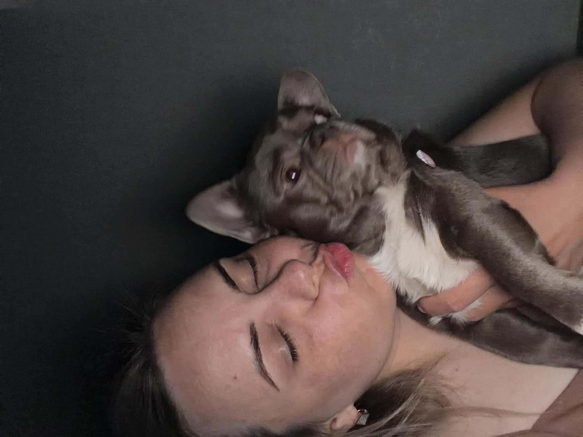 A woman is lying on a dark gray surface. She has her eyes closed and lips pursed as if she is kissing. A French Bulldog puppy, primarily gray with white chest, is being held close to the woman's face. The dog is looking upwards with one ear slightly bent. The woman is wearing a stud earring. Location is not specified in the image or its data.