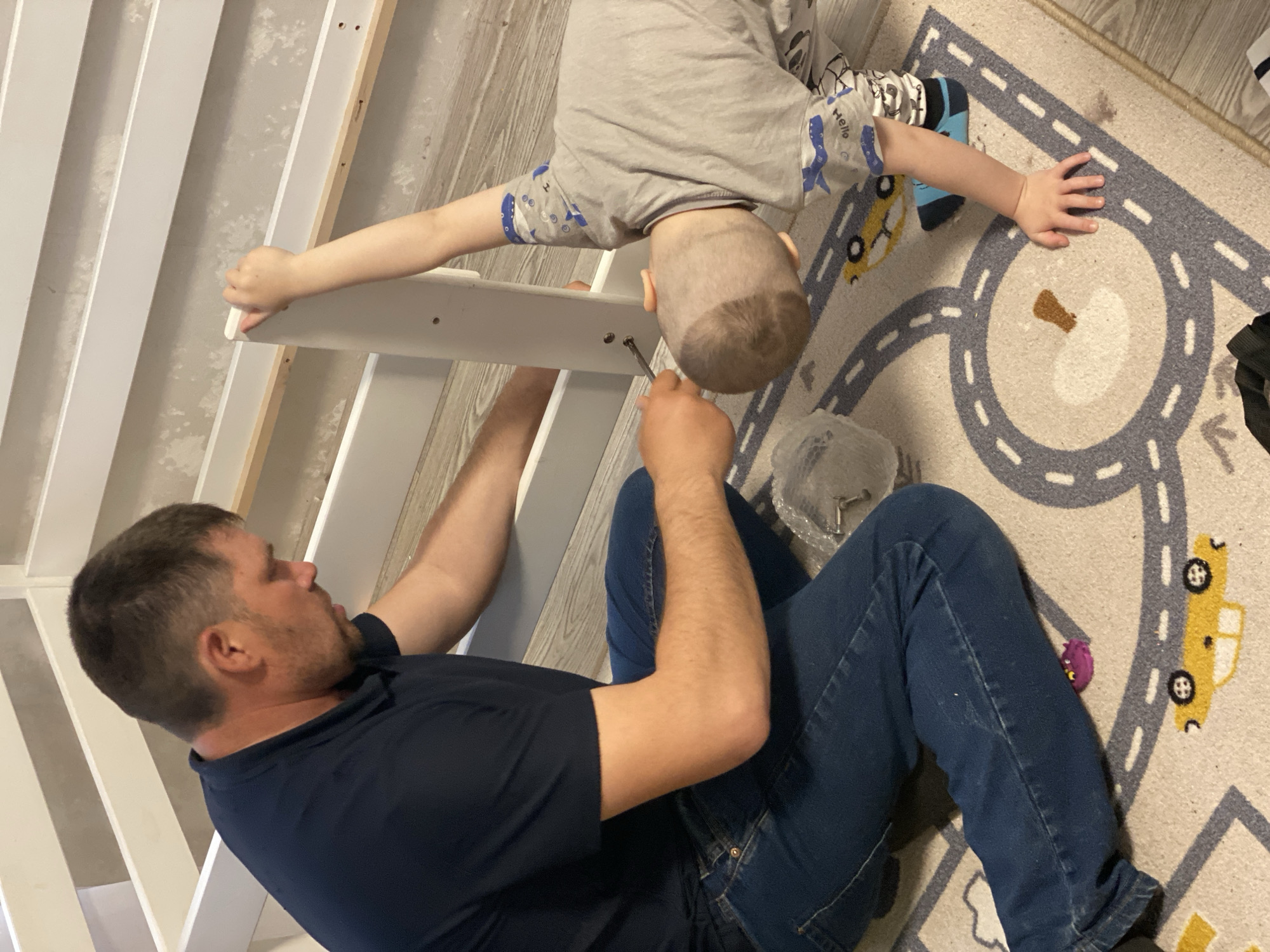 A man and a child assemble a white crib or bed. The man, wearing a navy blue shirt and blue jeans, uses a tool, possibly a screwdriver, to attach a piece of white wood. The child, in a grey shirt with blue pattern sleeves and shorts, assists by holding the wood piece. A bag of hardware sits nearby. They are on a carpet with a road design, including cars and paths. The location of Sochi, Russia, is not confirmed by visual evidence.