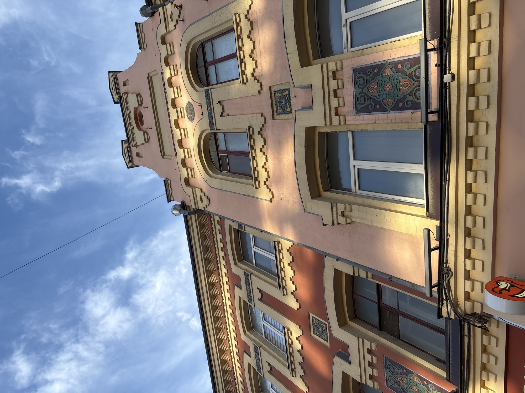 The image shows the facade of a building with a distinctive architectural style. The building is painted in two main colors: a light pink and a reddish-brown, with cream-colored decorative elements. The facade features arched windows with dark frames, as well as rectangular windows with white frames. There are also decorative tiled panels with intricate green and red patterns. The architectural style suggests an older building, possibly from the late 19th or early 20th century.
The sky is blue with scattered white clouds, indicating daylight. The sunlight casts shadows on the building, highlighting the textured decorative elements and suggesting it is either morning or late afternoon.
A thin black wire runs diagonally across the sky, attached to a light fixture on the building. Electrical cables are also visible running vertically along the side of the building. A small round sign with a stylized orange chicken logo is attached to the building on the right side.
The orientation of the image is such that the building appears to be viewed from a low angle, looking upwards towards the sky. The scene is an outdoor urban environment.
No people are visible in the image. The overall mood is one of architectural appreciation and a snapshot of a street view.
The location context provided is Novaya Usman', Russia, which helps to place the scene geographically. There is no visible text in the image apart from the logo on the sign.