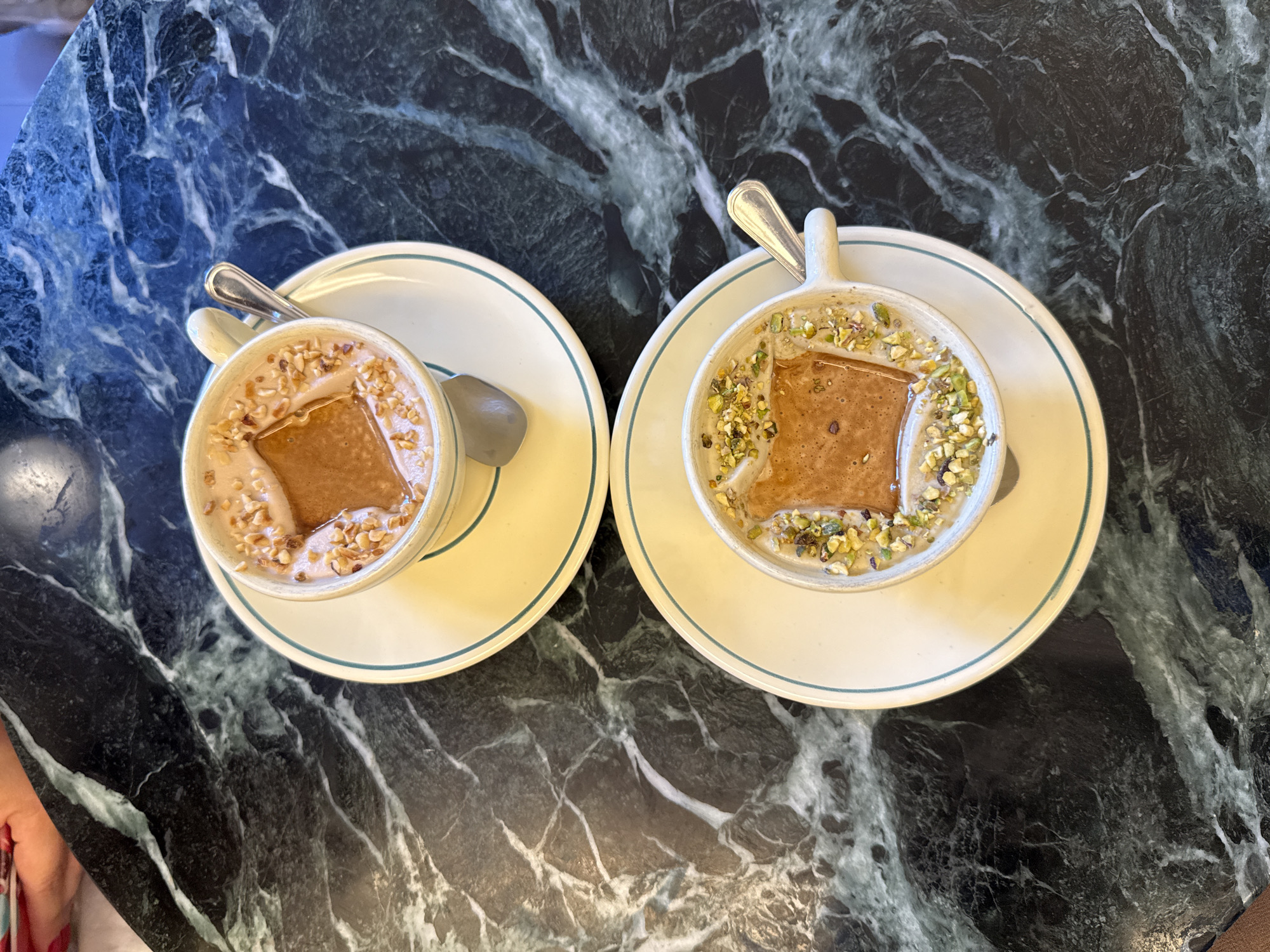 An overhead perspective captures two ceramic cups, each containing a creamy dessert or thick beverage, presented on a dark green marble table with prominent white and light green veining, characteristic of a cafe in Milan, Italy. Each white ceramic cup, featuring a handle, rests on a matching saucer detailed with a light blue rim. A silver spoon is positioned within each cup. The upper cup's contents are garnished with finely chopped brown nuts, identified as hazelnuts, and a rectangular light brown biscuit is partially submerged. The lower cup holds a similar creamy substance, garnished with finely chopped green pistachios, also containing an identical rectangular light brown biscuit. A blurred human hand is partially visible in the upper left quadrant of the image. The background is out of focus, indicating an indoor setting.
