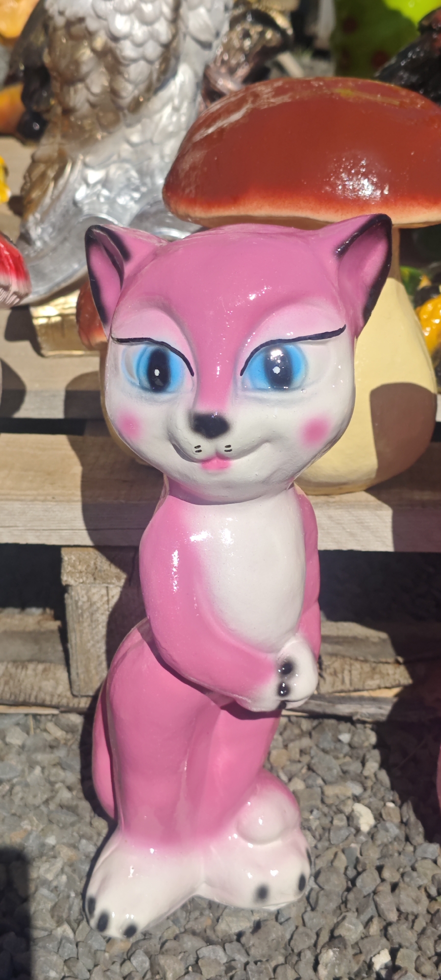 A pink ceramic cat figurine stands on gray gravel in Olen'e, Russia. The cat has white on its chest and paws, blue eyes, black whiskers, and pink cheeks. Behind the cat is a wooden plank, an owl figurine, and a mushroom decoration. The image is well-lit.