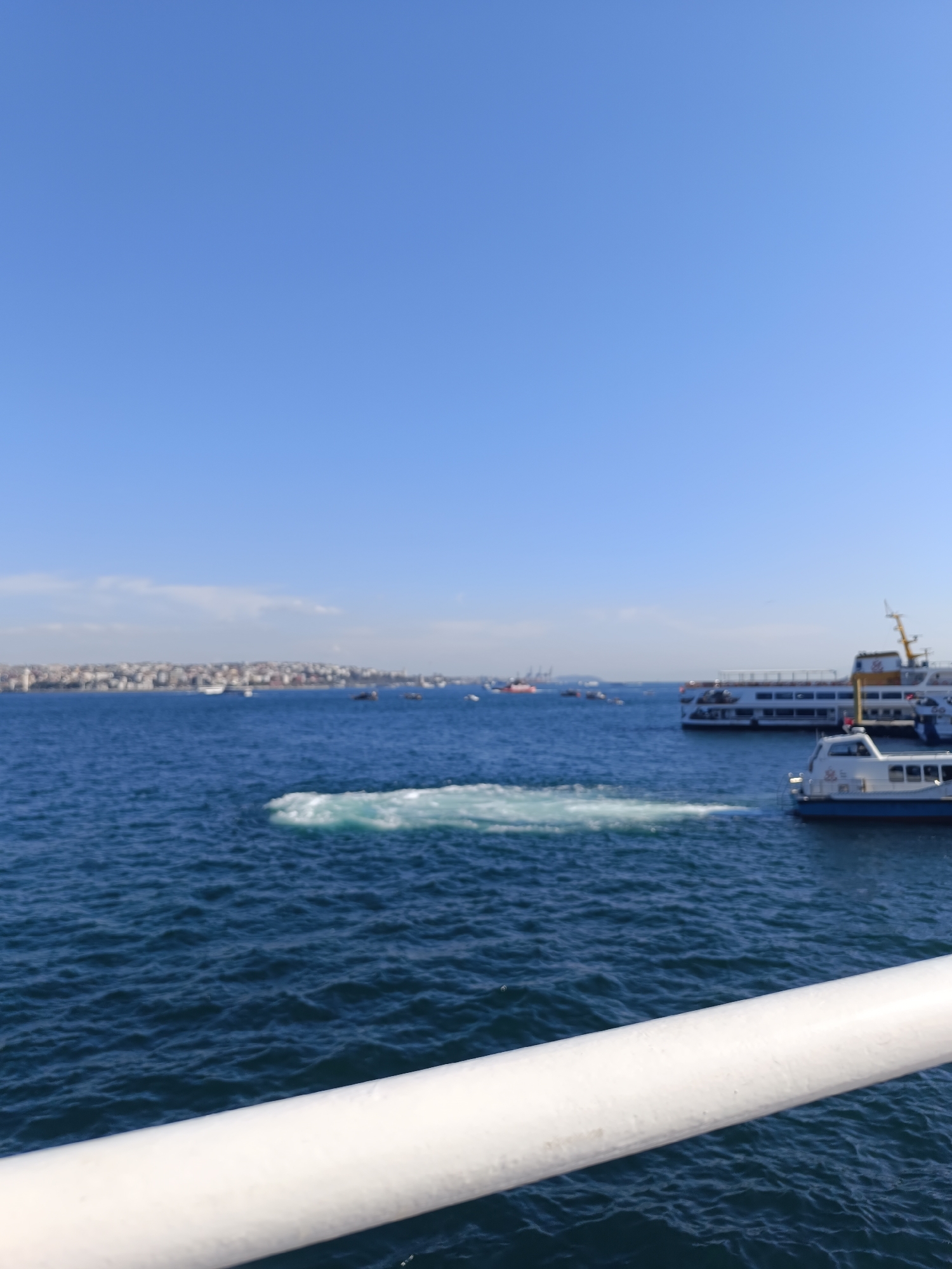 The image depicts a body of water with a white rail in the foreground. In the mid-ground, there are several boats and a landmass with buildings visible in the distance. The sky is clear and blue. The location appears to be a waterway in or near Beyoğlu, Turkey, given the architectural style of the buildings on the landmass. There's disturbance on the water's surface, potentially from a boat's wake or other activity.