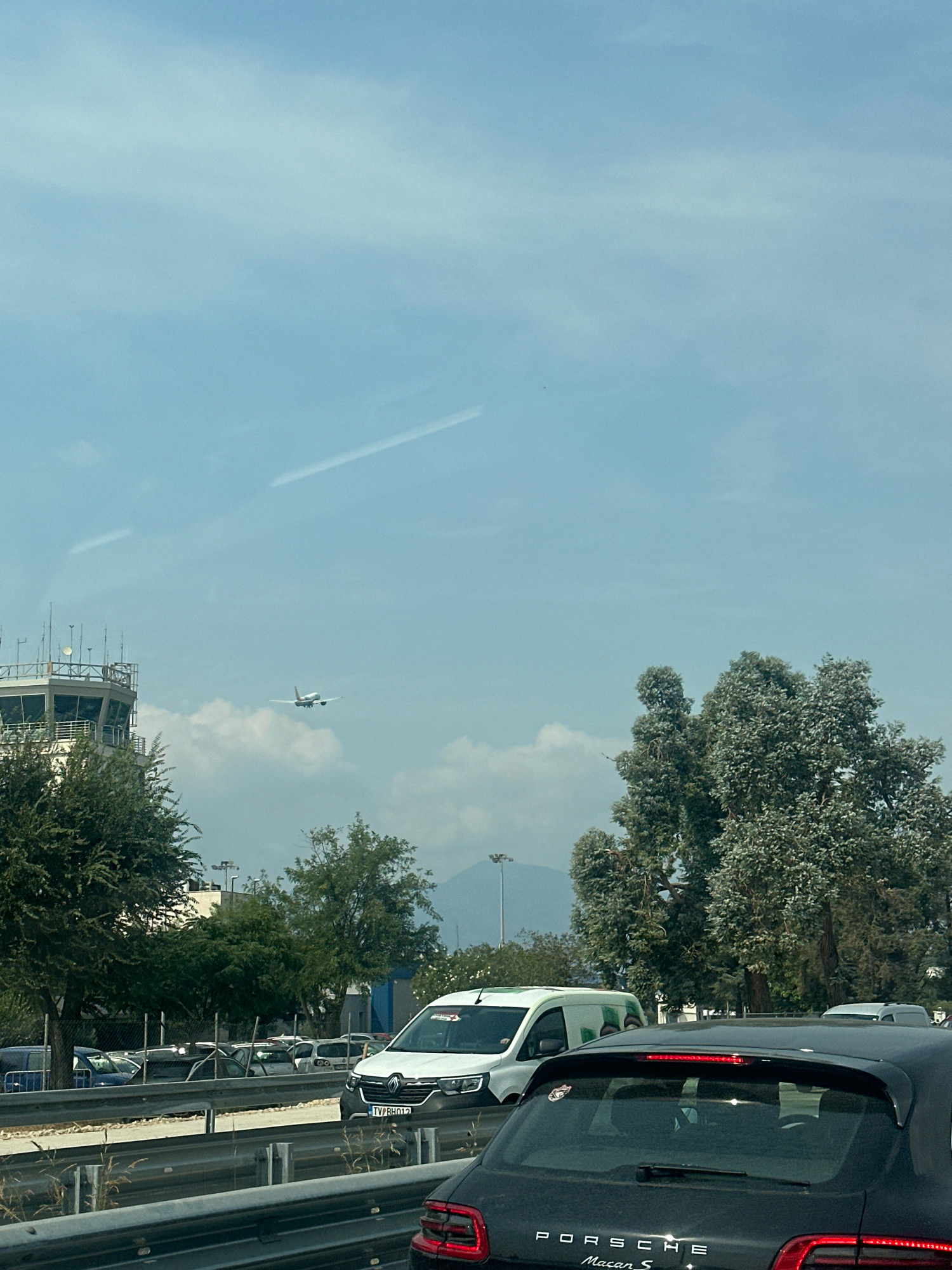 The image captures a scene on a highway likely near an airport, as suggested by the presence of an air traffic control tower and a plane in flight. The sky is a light blue with wispy clouds and a faint contrail from another aircraft. In the foreground, several vehicles are visible, including a black Porsche Macan S, a white van, and other cars in traffic. The air traffic control tower, a prominent structure with multiple antennas, stands to the left, partially obscured by trees. An airplane is ascending or descending against a backdrop of clouds. Trees and other greenery line the side of the highway, with some industrial-looking buildings in the distance. The license plate on the white van reads "TV PR 012". The overall impression is of a typical traffic situation on a sunny day near an airport. The context provided indicates the location is Mrčevac, Montenegro, likely near Tivat Airport.