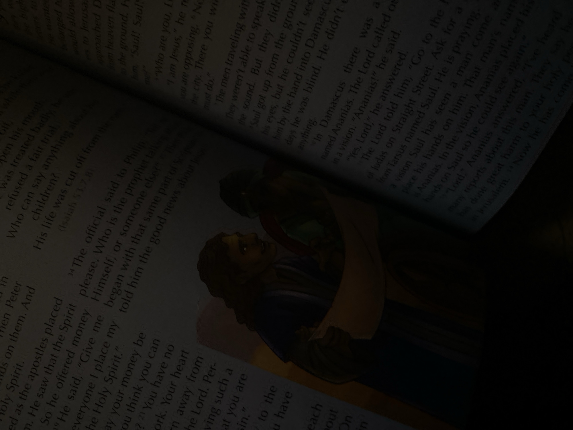 A close-up, dimly lit photograph shows an open book, with text visible on both pages. The right page features a watercolor illustration of two figures standing over a document. The left page displays a passage from the Bible, with verses numbered. The lighting suggests the book is illuminated by a lamp or natural light source that creates a warm glow on the illustration and text. The image is framed such that the bottom portion is dark, obscuring any further context. There is no visible indication of Jauro Yinu, Nigeria, in the image.