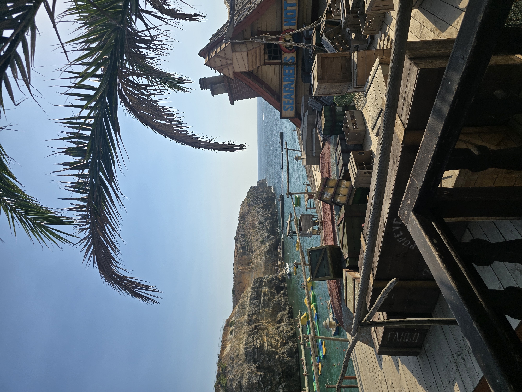 The image depicts a scenic view of Popeye Village, a film set and tourist attraction located in the city of Mellieħa, Malta. In the foreground, wooden crates are stacked on a wooden platform. A weathered wooden railing is also visible in the foreground, obscuring some of the crates. In the mid-ground, a rustic, wooden building with a red roof is adorned with a sign that reads "SEAFARERS HOTEL." The building has a chimney and windows. More wooden crates are scattered around the building. The background features a picturesque bay with clear turquoise water. Numerous inflatable water toys are floating in the water, and several boats are anchored or in motion. Rocky cliffs rise majestically on the left side of the bay, and people can be seen swimming and relaxing on platforms in the water. A palm frond hangs from the top of the frame, partially obscuring the bright blue sky. The overall scene evokes a sense of a charming, Mediterranean coastal village, likely set up to resemble a pirate or sailor's haven.