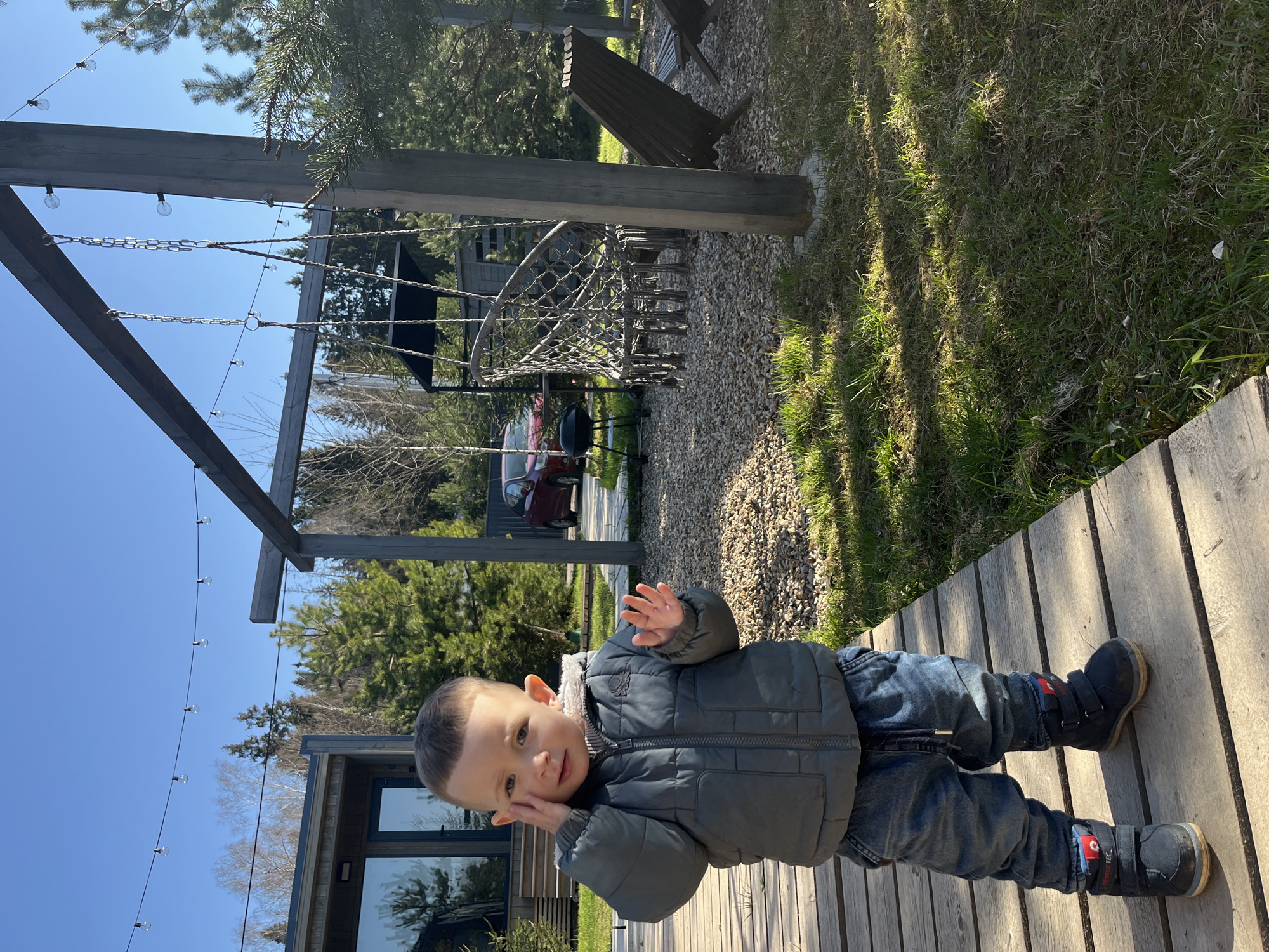 A toddler is standing on a wooden deck. The child is wearing a grey jacket, jeans, and black boots with red accents. The deck is made of light-colored wooden planks. In the background, there is a playground structure with wooden beams, chains, and a climbing net. Beyond the playground, there are trees, grass, and a building with large windows. A red car is parked nearby. The sky is blue. The location is Литвиново, Russia.
