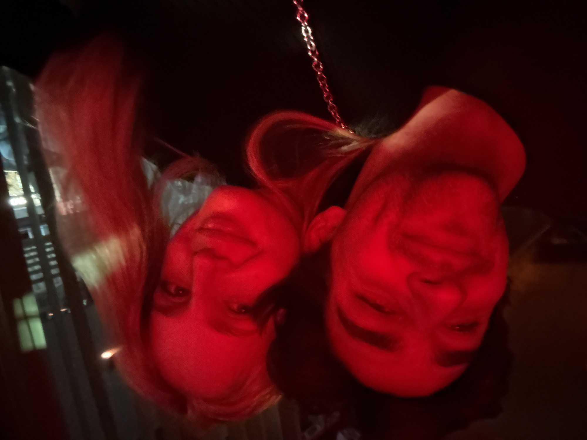 Two people are in the image. The person on the left has long hair and is smiling, while the person on the right appears to have short hair and is also smiling. The image is heavily lit in red and appears to be taken at night. A metal chain is visible. The location is unknown and cannot be determined with the provided image.
