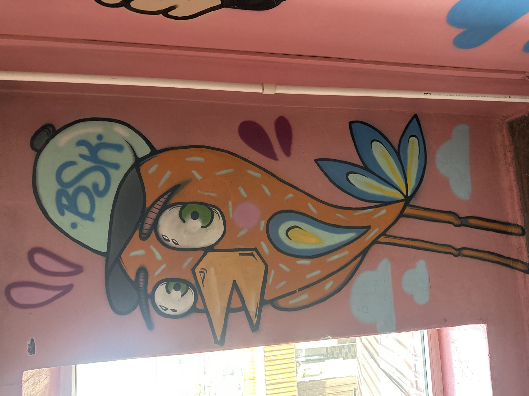 This is a close-up, eye-level, vertical photograph of a colorful graffiti mural on a pink wall. The mural depicts a cartoonish orange bird wearing a light blue baseball cap with the letters "BSK" written in blue on it. The bird has large, wide-open eyes with green irises, an orange beak, and small, light blue clouds are painted around it. The bird has a decorative pattern of thin, light blue dashes across its body. Its legs are long and thin, painted orange. In the upper left corner, there are two pink shapes resembling bunny ears. To the right of the bird mural, a white pipe runs vertically along the wall, and a portion of another graffiti element, possibly a blue heart, is visible on the far right. The background wall is solid pink, with a window to the left showing some indistinct yellow structures and a slightly overcast sky. The photograph appears to be taken indoors, likely in an alley or hallway. The graffiti art is done in a bold, outlined style with vibrant colors. This location is identified as Biysk, Russia.