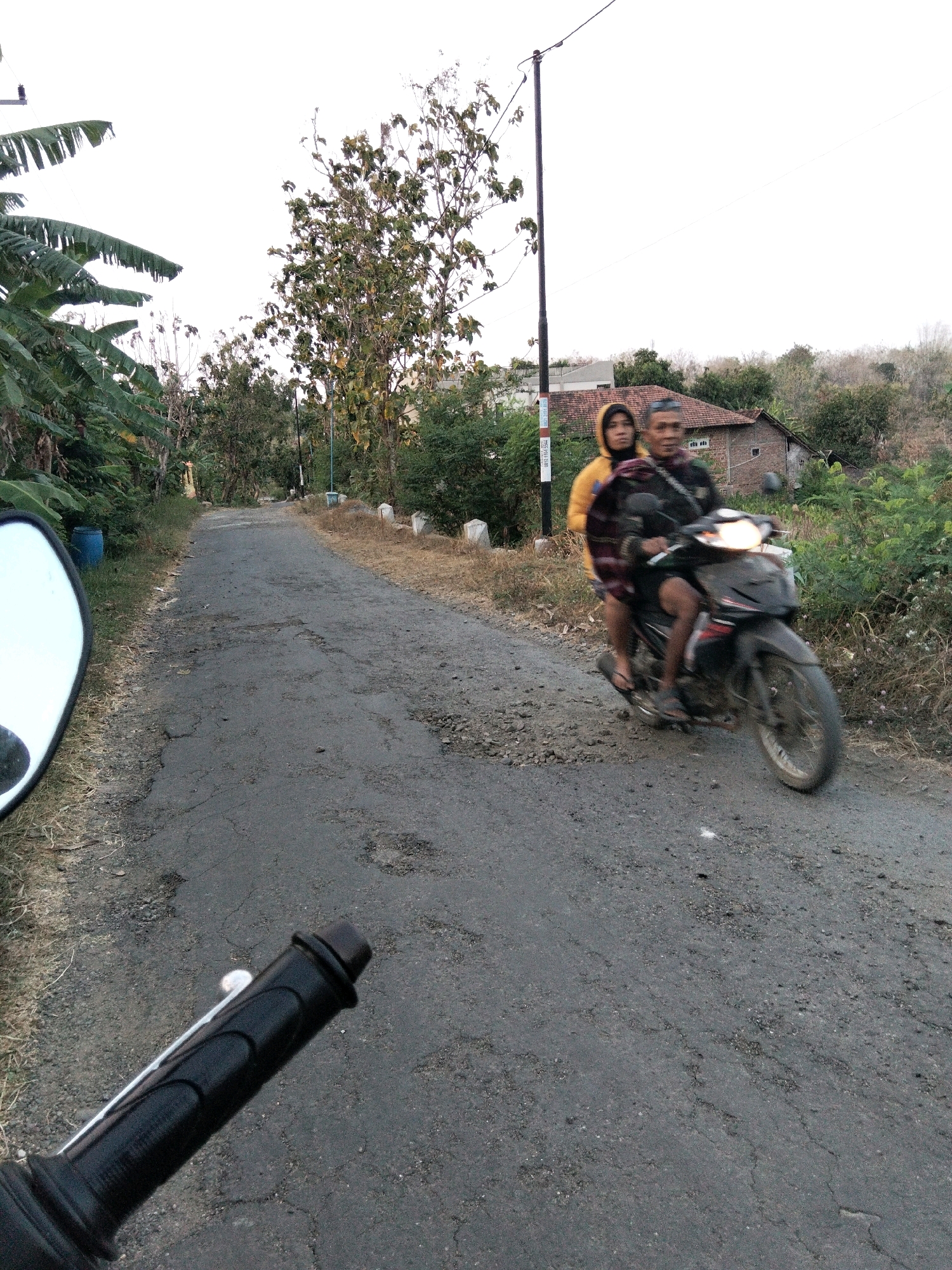 A man and a woman are riding a motorcycle on a cracked asphalt road in a rural area. The woman is wearing a yellow hoodie and the man is wearing a dark shirt. They are both looking forward. The road is lined with trees and bushes. In the background, there is a house with a red tile roof and a few other buildings. The sky is overcast. The photo appears to have been taken in the morning or late afternoon. The location is Jenangan, Indonesia.