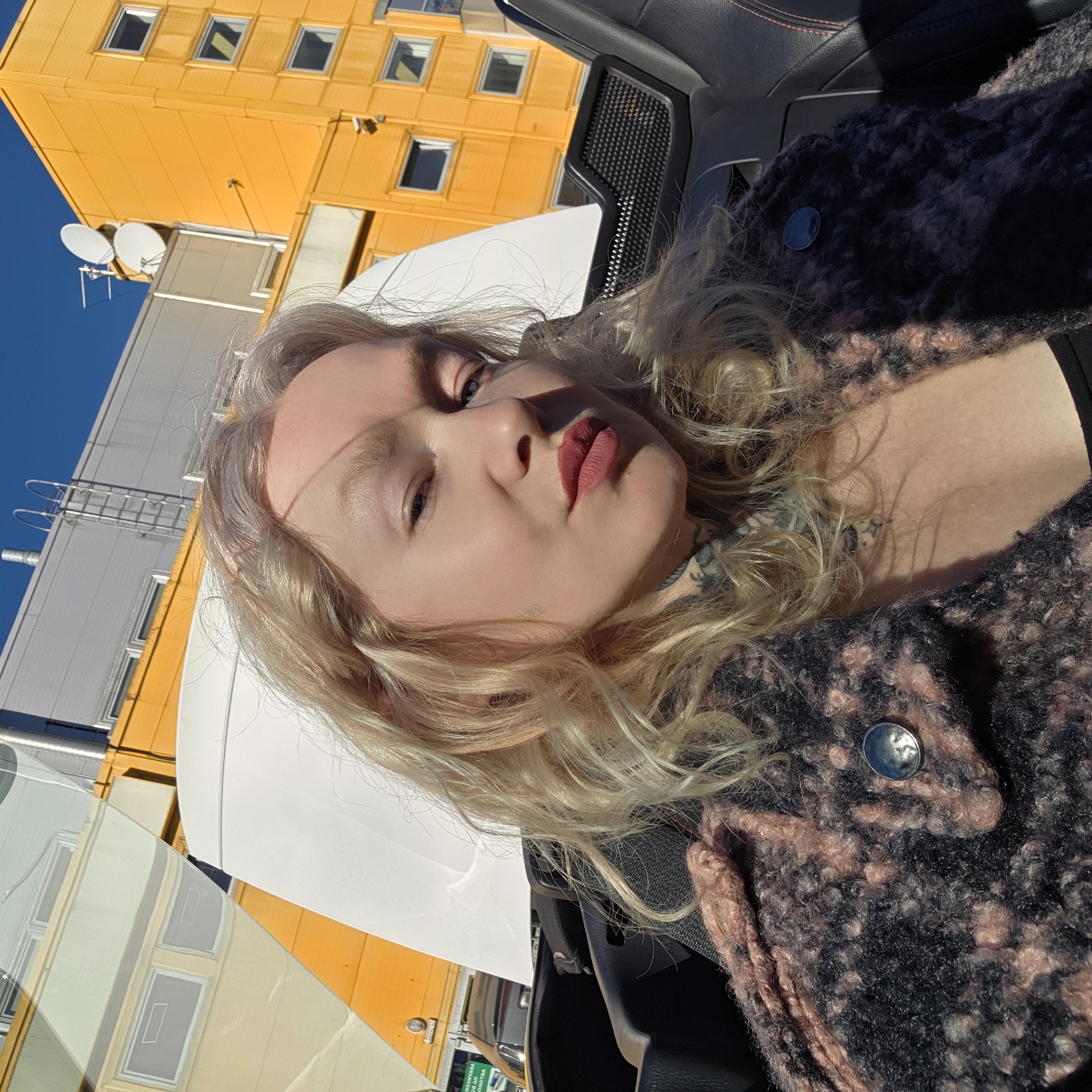 A blonde woman with fair skin is posing for a selfie in the driver's seat of a car. She is wearing a patterned coat or jacket with a button closure. Outside the car window, there are several buildings, including a yellow building with many windows and a silver-colored building with a ladder attached to the side. The sky is blue and sunny. Based on the architecture, color scheme of buildings, the types of vehicles, and general look, this photograph was most likely taken in Saint Petersburg, Russia.