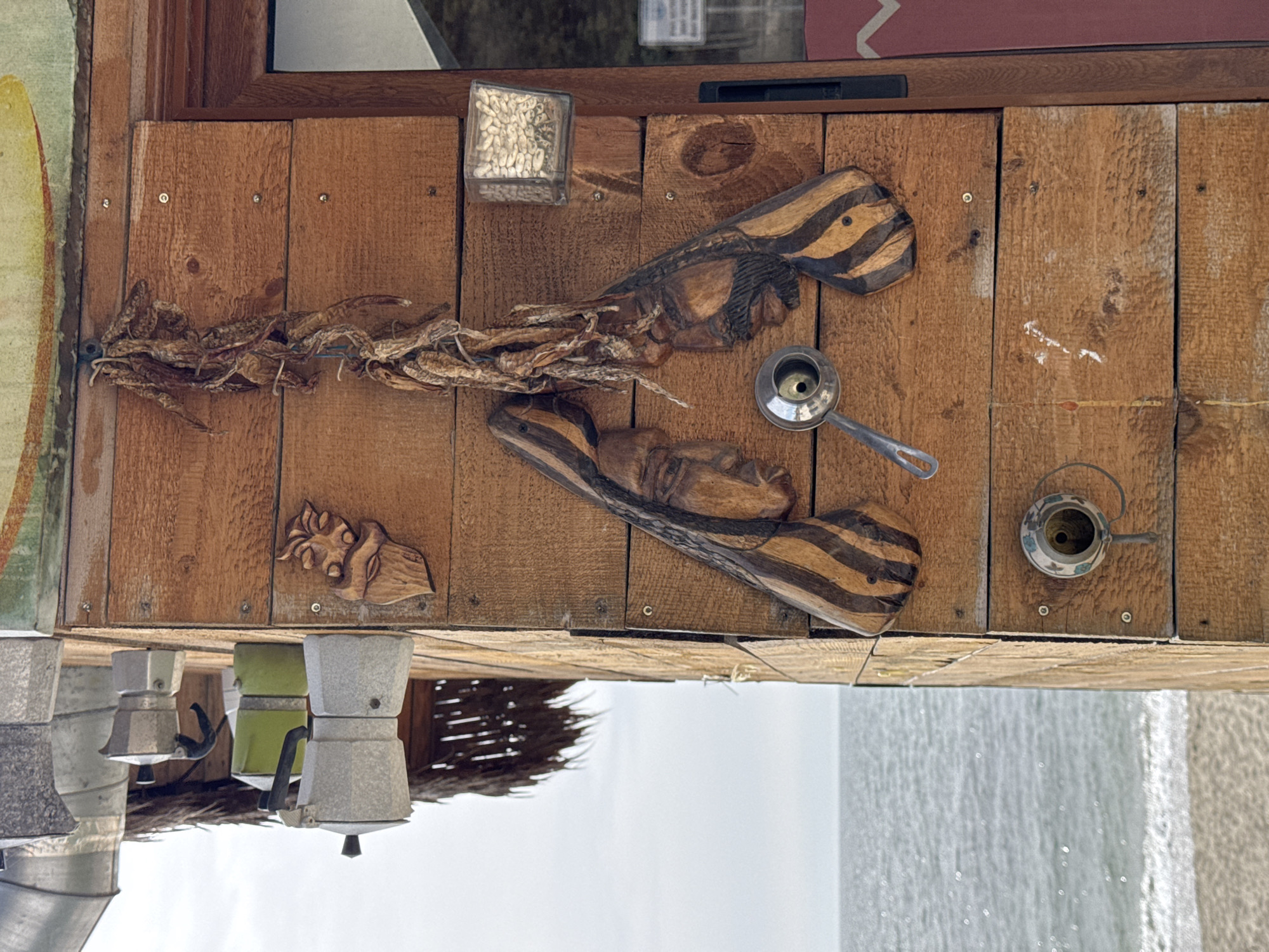 The image shows a detailed section of a wooden structure in Varna, Bulgaria, possibly a storefront or an outdoor café, mounted on a building facade. The wooden plank wall is adorned with various decorative items. On the left is a string of dried chili peppers, next to a clear cube containing white beans or seeds. There are two carved wooden pieces with alternating dark and light stripes, appearing as stylized human heads, placed above each other. A small, shiny, metal scoop or measuring spoon is positioned diagonally to the upper right of the lower carving. Towards the right of the wooden plank wall, a small, decorative teapot is attached.
Below the wooden structure, several Italian Moka pots are visible, suggesting the preparation or sale of coffee. The exterior wall shows a weathered, possibly plastered or textured finish. A partial view shows a pale sky.