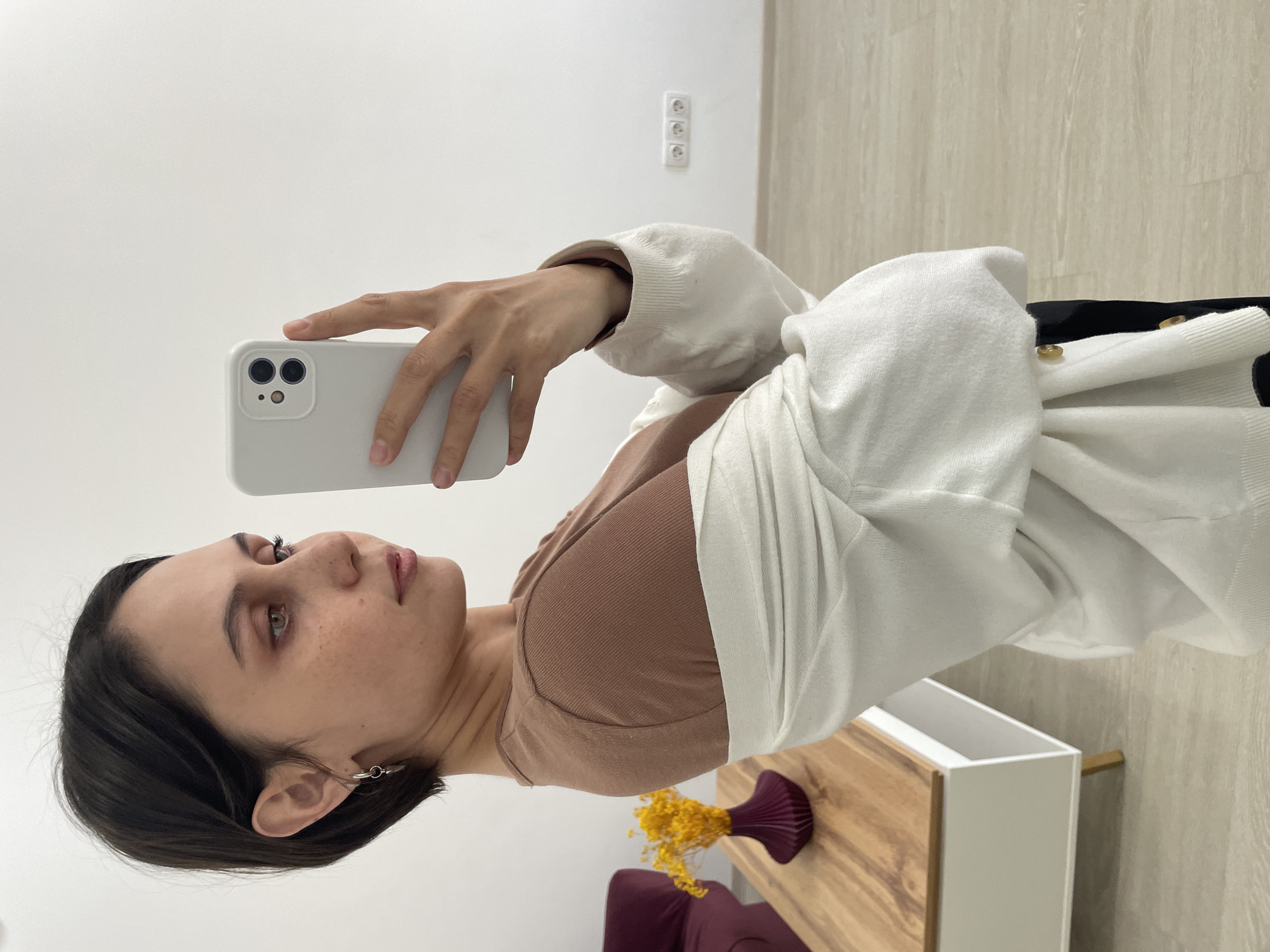 This image, captured indoors in what appears to be a modern room in Izhevsk, Russia, depicts a young woman taking a selfie. The photograph is vertically oriented, with the subject positioned on the left.
The woman has dark hair, light-colored eyes, and some freckles on her face. She is wearing a light brown or beige ribbed top underneath an open, white or off-white cardigan, which has small golden-colored buttons. A simple silver hoop earring is visible in her left ear. She holds a light-colored smartphone, identifiable by its dual-camera module, in her right hand, extended to take the selfie. Her expression is neutral and focused on her phone.
The background features a plain white wall. On the wall, a white electrical outlet with three round sockets is prominently visible, characteristic of European Type F or E sockets used in Russia. Below this, a light wooden piece of furniture, possibly a side table or console, features a white, open compartment. Adorning the wooden surface is a distinctive dark purple, pleated vase containing bright yellow dried flowers, possibly baby's breath. A glimpse of a dark red or burgundy upholstered chair or sofa is seen in the lower left corner. The floor is a light-toned wood or laminate. The bright, even lighting suggests it is daytime. There is no visible text in the image.
