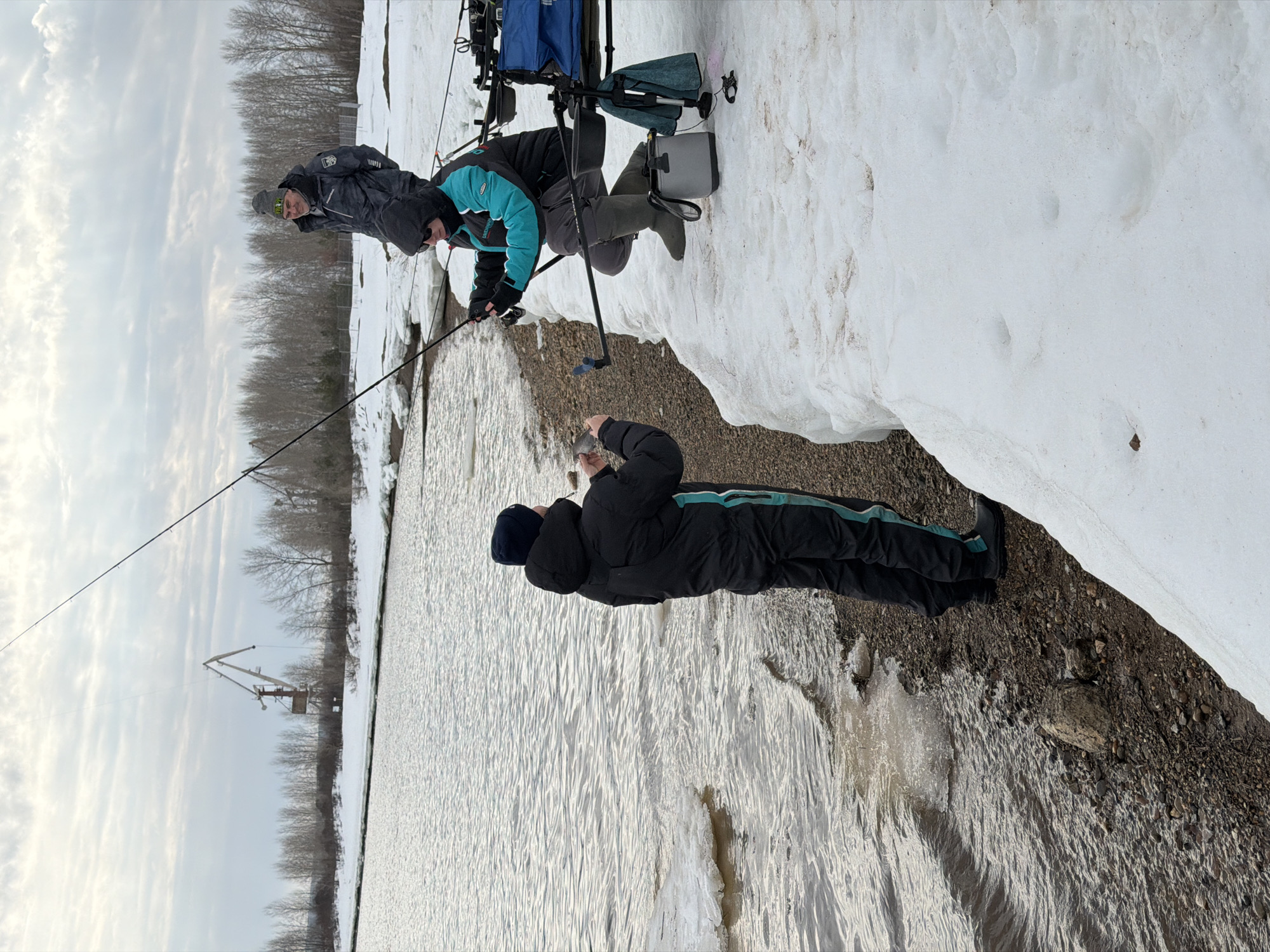 The image depicts two individuals ice fishing in Noviy Urengoy, Russia, amidst a winter scene. One person, clad in a black jacket with a teal stripe, lies prone on the snow-covered bank, holding what appears to be a fish in their hand. A second person, wearing a gray jacket and teal gloves, leans over the edge of the snow-covered bank towards the water with a fishing rod. Fishing equipment, including a chair and tackle box, is situated on the snowy ground behind the second person. The water is partially frozen, and a tall industrial crane is visible in the background against a cloudy sky. The overall scene suggests a cold, outdoor environment with active participation in the recreational activity of ice fishing.