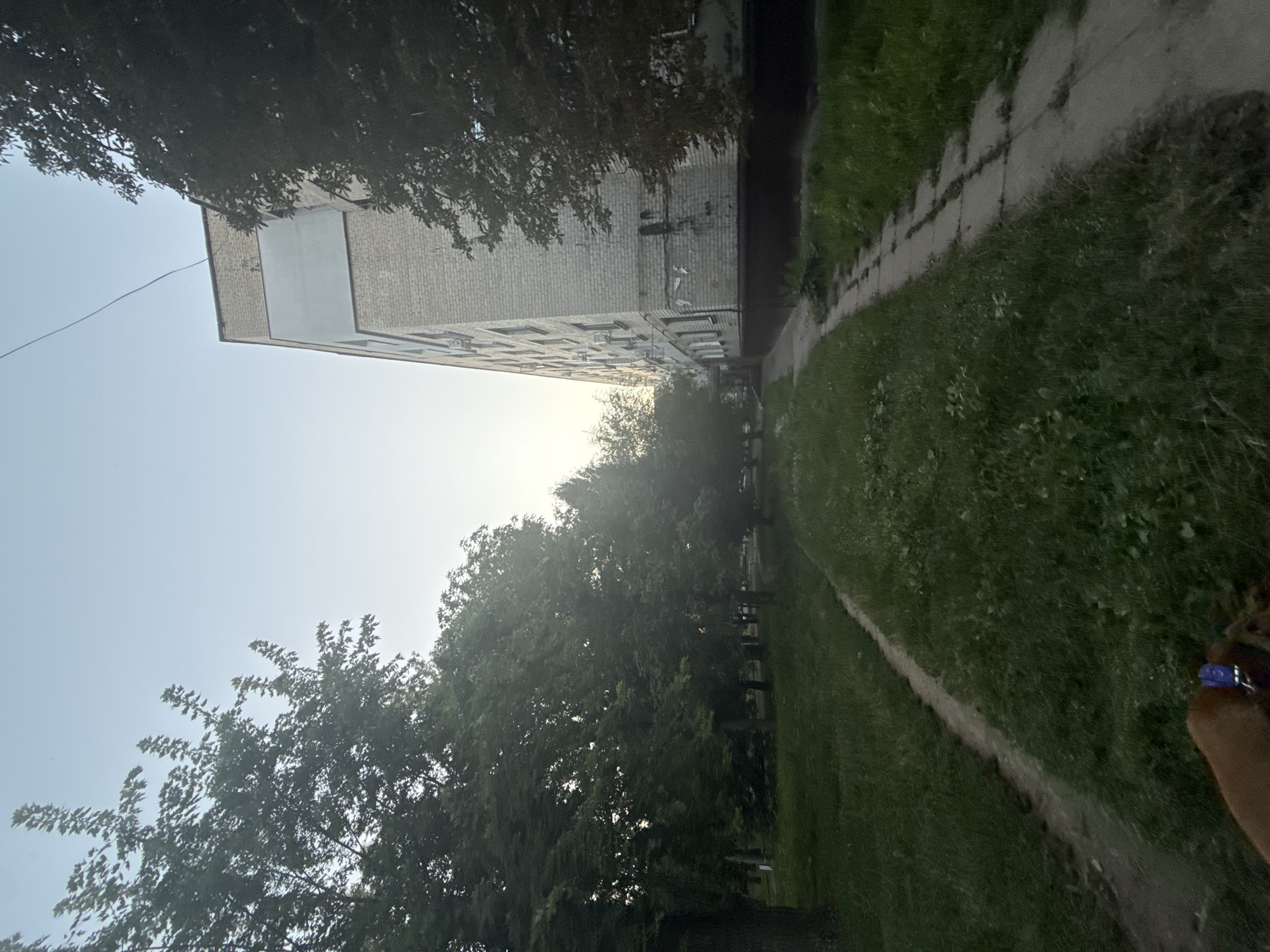 This image captures a view of a multi-story apartment building in a residential area, likely in Zaporizhzhia, Ukraine, given the provided context. The building, constructed from light-colored bricks, features multiple windows and air conditioning units mounted on its facade. The time of day appears to be either early morning or late evening, as indicated by the soft, diffused light and the clear sky.
In the foreground, a narrow, unpaved path runs alongside a concrete walkway, bordered by lush green grass. Trees with dense foliage line the left side of the scene, casting shadows and creating a natural canopy. The overall environment suggests a typical urban setting with green spaces integrated into the residential landscape. A part of a dog's head and harness is visible in the bottom left corner of the frame, suggesting it is being walked.