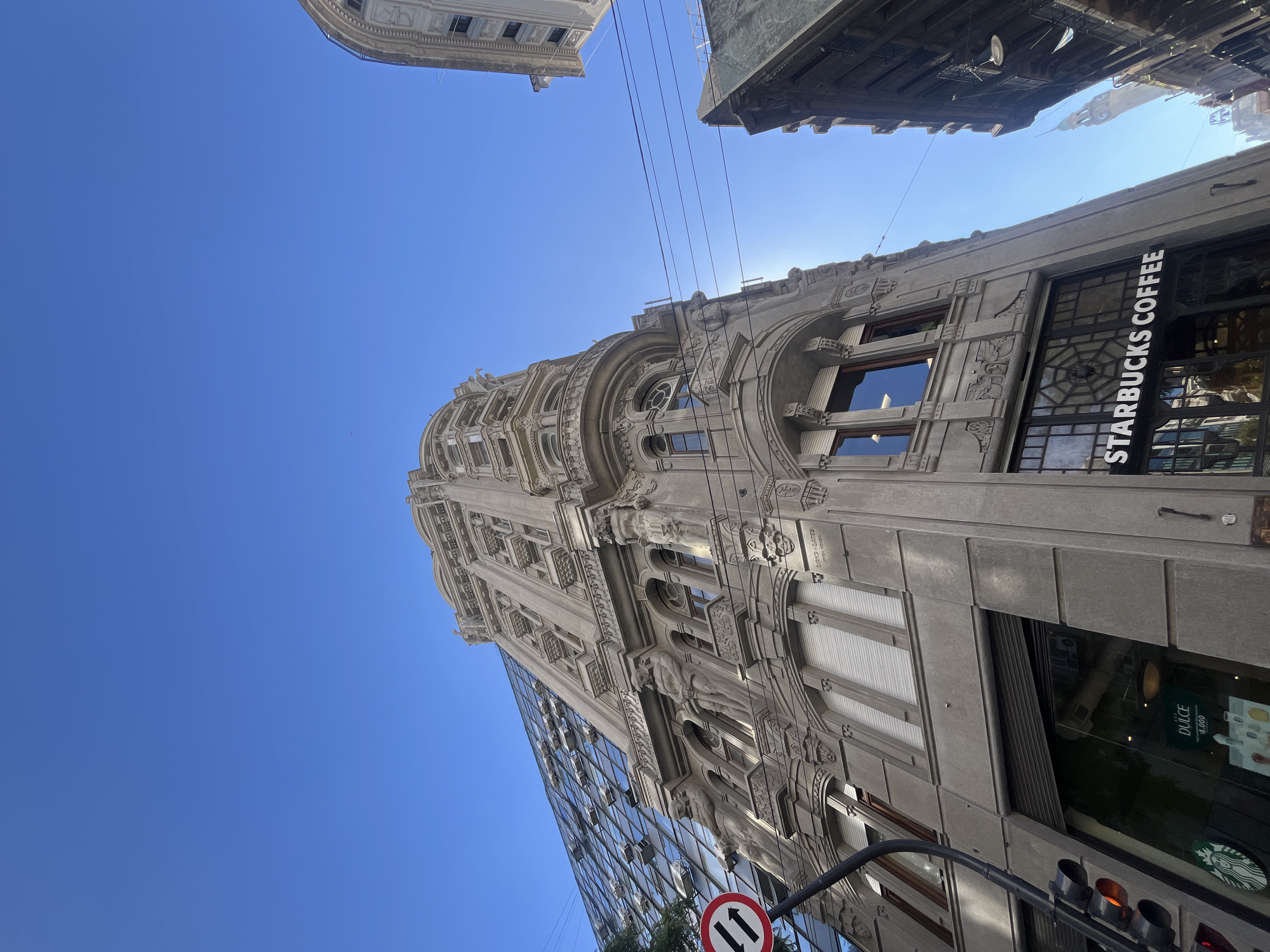 The image presents an upward-angled view of an urban street scene in Buenos Aires, Argentina, bathed in bright daylight under a clear blue sky. The primary subject is a tall, ornate building with a stone or concrete facade, characterized by rich architectural details, including elaborate carvings, decorative balconies, and arched windows. The sun is visible behind the upper section of this building, creating a halo effect.
Adjacent to this historic-looking structure, a modern building with a sleek glass facade reflects the sky and surroundings. Overhead, several power lines crisscross the blue sky.
Visible text includes "STARBUCKS COFFEE" displayed vertically on a large window of the ornate building. Inside this window, a sign reads "DULCE" with additional smaller text, possibly "SABORES UNICOS" and "SIEMPRE SABES." The distinctive green Starbucks logo is also visible. At the bottom of the frame, a black traffic light pole with red and amber lights is partially visible, alongside a circular road sign with a red border, white background, and two black vertical arrows, indicating two-way traffic. Another building can be seen in the upper left background, and the corner of another structure is visible in the upper right. The scene depicts a lively city environment with a blend of old and new architectural styles.