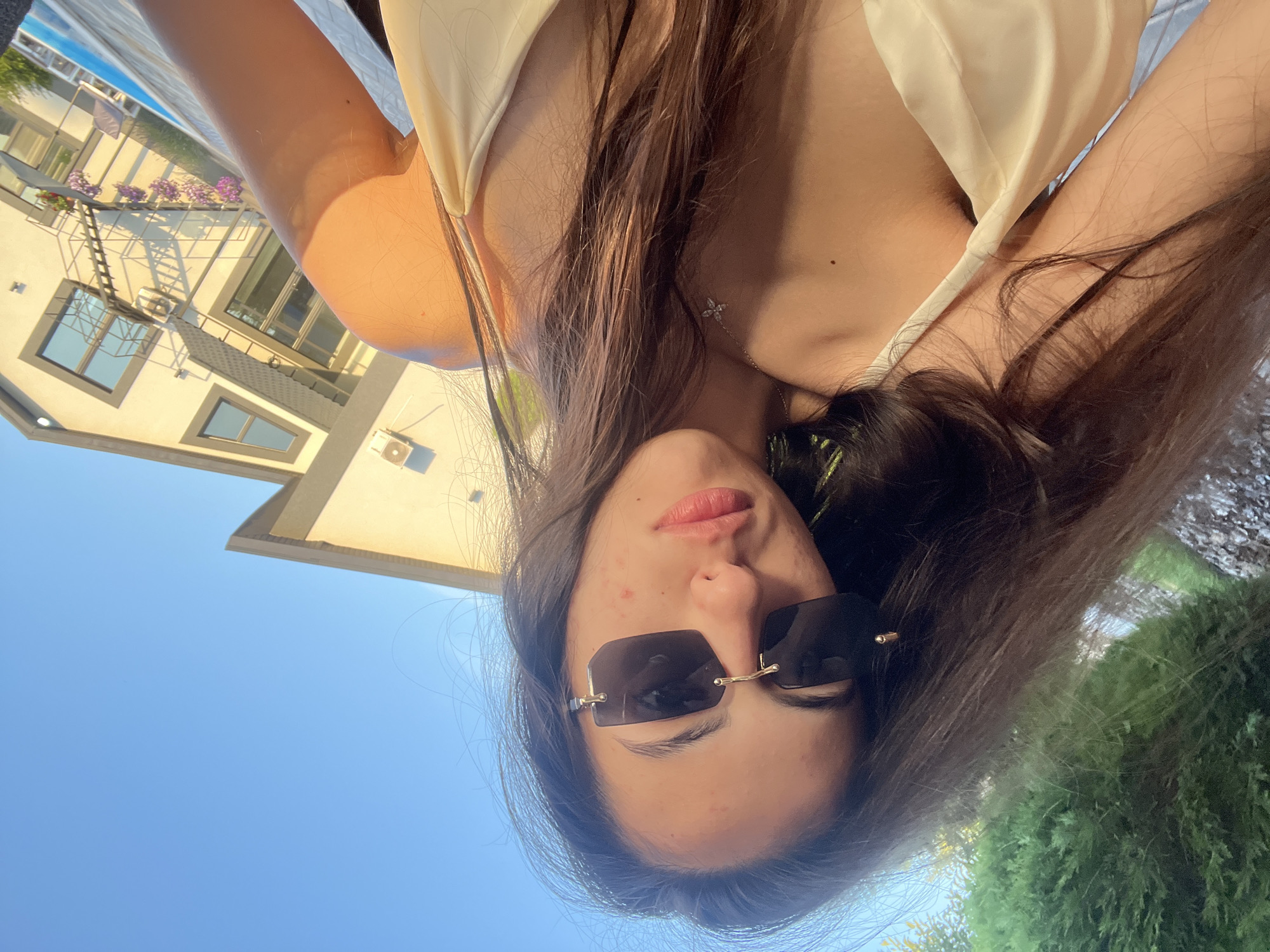 The image features a young woman taking a selfie outdoors. She has long, dark brown hair, is wearing sunglasses, and a light-colored top. Her face is the primary focus, and she appears to be looking towards the camera. She has some minor blemishes on her skin.
The background is a bright blue sky, suggesting it is daytime and the weather is clear. To the left of the frame, part of a building is visible, with windows and a ladder leading up to a balcony. There are also some potted flowers on the balcony. To the right, there is a lush green hedge.
The orientation of the image is rotated, making it appear as if the subject is lying down or the camera is held upside down. This perspective, combined with the outdoor setting, suggests a relaxed or casual moment, possibly by a pool or in a garden. No text is visible in the image. The location context provided is Novooleksandrivka, Ukraine.