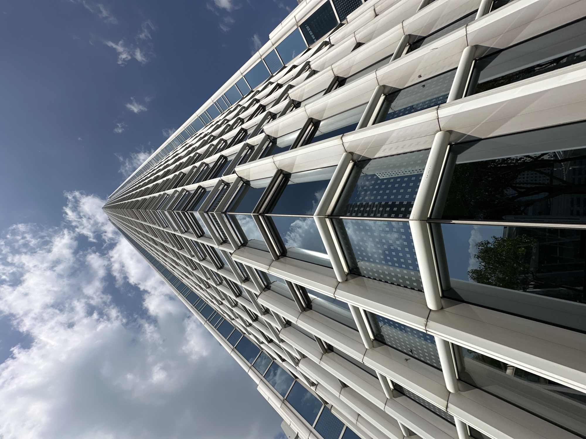 This is an upward-angled, low-angle shot of the International Finance Centre (IFC) skyscraper in Hong Kong, China. The building's facade is made of reflective glass windows and white mullions. The sky above is a deep blue with scattered white clouds. The reflections in the glass show the sky, clouds, and other buildings. The perspective creates a dramatic effect, emphasizing the height and scale of the skyscraper. There are no people visible in the image.