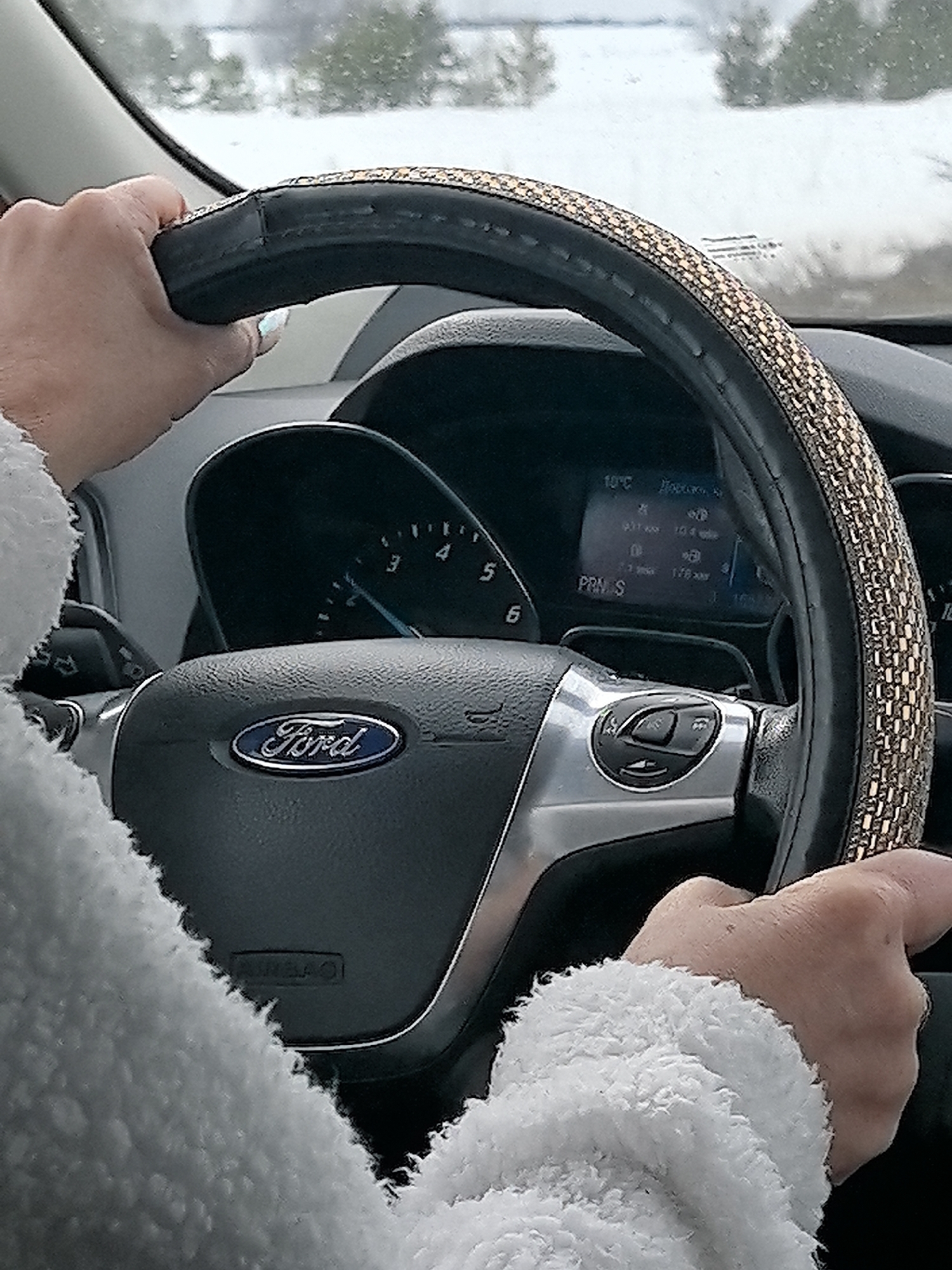 The scene is set inside a Ford vehicle, in Ledukhi, Russia, with a wintery exterior visible through the windshield. Two human hands, appearing to be a light skin tone, grip a black steering wheel, which is adorned with a textured, golden-brown decorative cover. The left hand is positioned on the upper-left quadrant, and the right hand on the upper-right quadrant. The driver wears a light-colored, fluffy, possibly fleece garment, visible on the forearms. The steering wheel prominently displays the blue oval Ford logo on its central airbag cover and features silver-toned control buttons on the left spoke. Behind the steering wheel, the instrument cluster is visible, including a partial view of an analog gauge on the left and a digital display showing "10°C", "PRN_S" (indicating current gear selection), and other Cyrillic text with numerical data, including "178 км". The exterior environment, seen beyond the dashboard, shows snow-covered ground and trees under what appears to be an overcast sky, indicating cold weather conditions.
