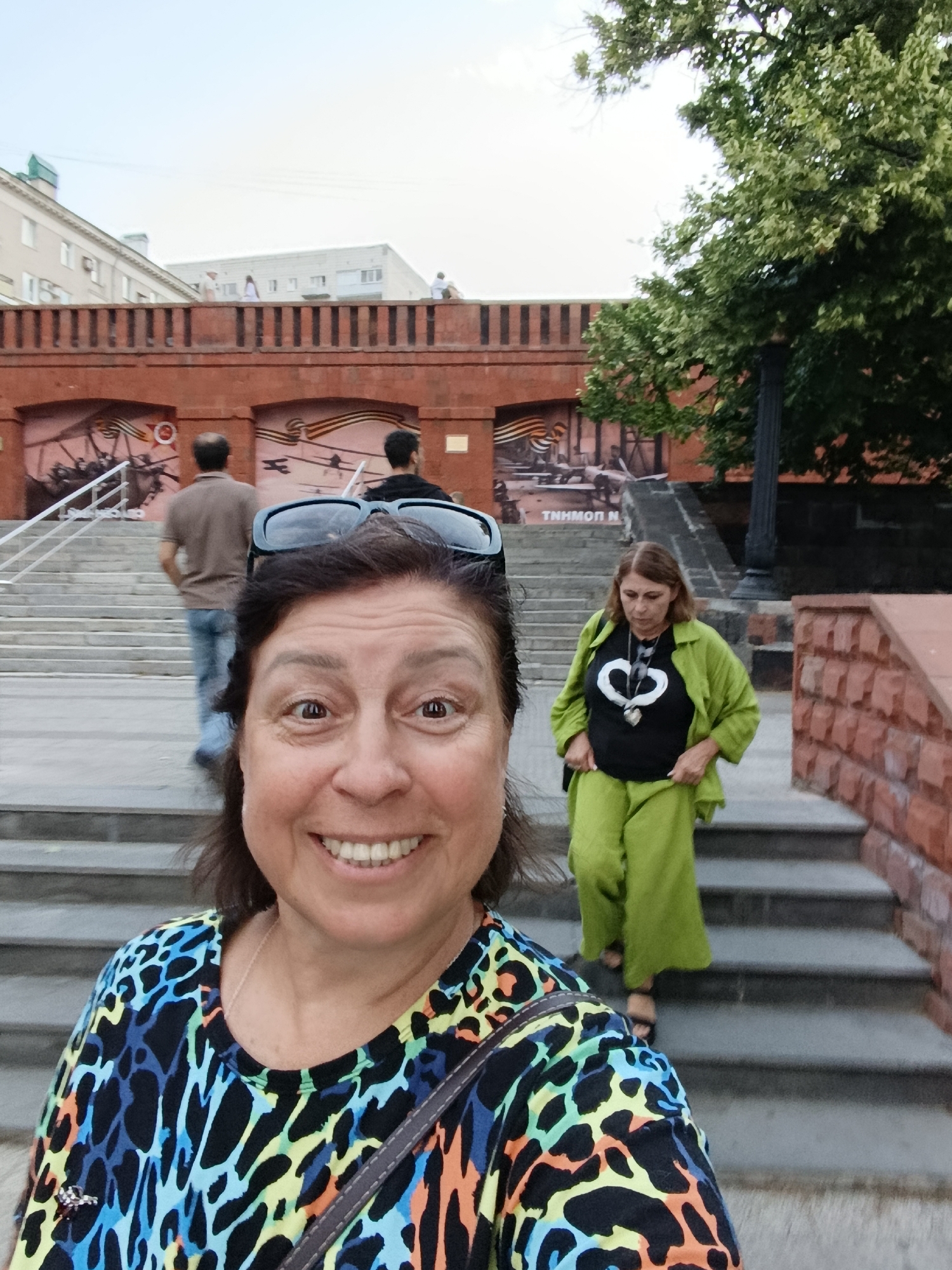 In this close-up selfie, a woman with dark hair and a broad smile is positioned in the foreground, looking directly at the camera. She is wearing a colorful, patterned top with leopard-like spots in black, blue, yellow, and orange. Sunglasses are perched on top of her head. Behind her, a woman in a bright green outfit consisting of a jacket and wide-leg pants walks down a set of concrete stairs. She is wearing a black t-shirt with a white heart design and a necklace with a pendant. Further in the background, two men are walking up the stairs. One man wears a brown t-shirt and jeans, while the other wears a dark t-shirt. Above the stairs, a red brick structure with murals depicting war scenes is visible, featuring planes and the St. George ribbon. Buildings and trees form the backdrop under an overcast sky.