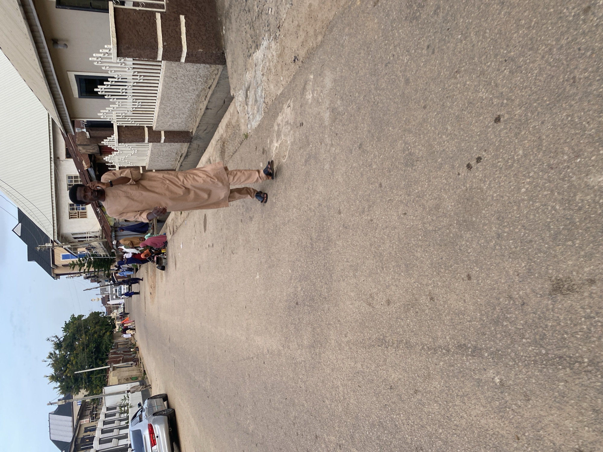 A man in a beige kaftan and trousers is walking on a paved street in Jos, Nigeria, while talking on his cell phone. He is wearing dark sandals. Several other people, some in traditional clothing, are visible in the background, some sitting by the side of the road, others standing. A silver car is parked on the left side of the street. Buildings line both sides of the street, some appearing to be residential homes with fences. The sky is overcast.
