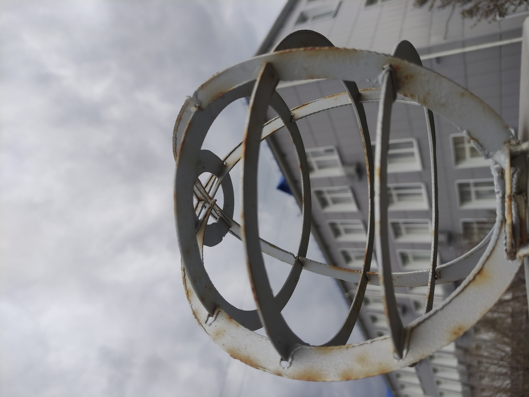 A low-angle shot shows a weathered, spherical metal structure with circular bands and vertical supports against a backdrop of a multi-story building and a cloudy sky. Rust is visible on the metal frame. The building features rows of rectangular windows and a neutral color palette. Patches of vegetation are seen at the bottom right corner of the frame. The structure is presumably located outdoors.