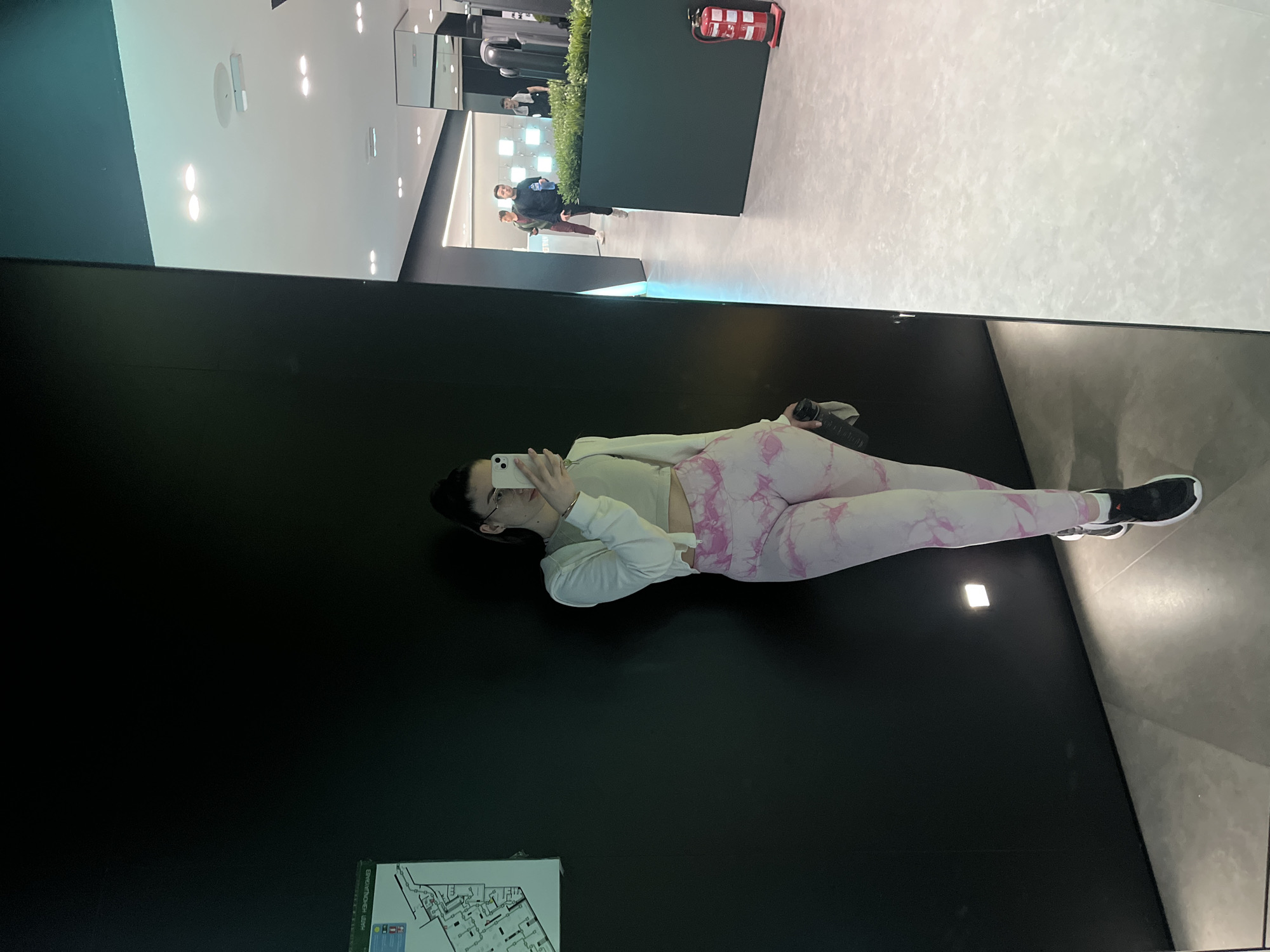 A vertically-oriented image shows a woman lying on a black surface, holding a white phone in her left hand. She has dark hair and wears glasses, a light-colored shirt, a white hoodie, and pink marble-patterned leggings. She has black shoes with white soles on her feet. To her left is a white sign with dark writing and diagrams. In the background, out of focus and reflected in what appears to be a large mirror or window, is a gym with equipment and other people. The room has a white ceiling and is well-lit with recessed lighting. There is a dark green wall with plants behind it. An object resembling a red fire extinguisher is on the wall. Two men are visible in the background, one wearing a black sweater. The location is a gym. The city and other detailed information are unknown.