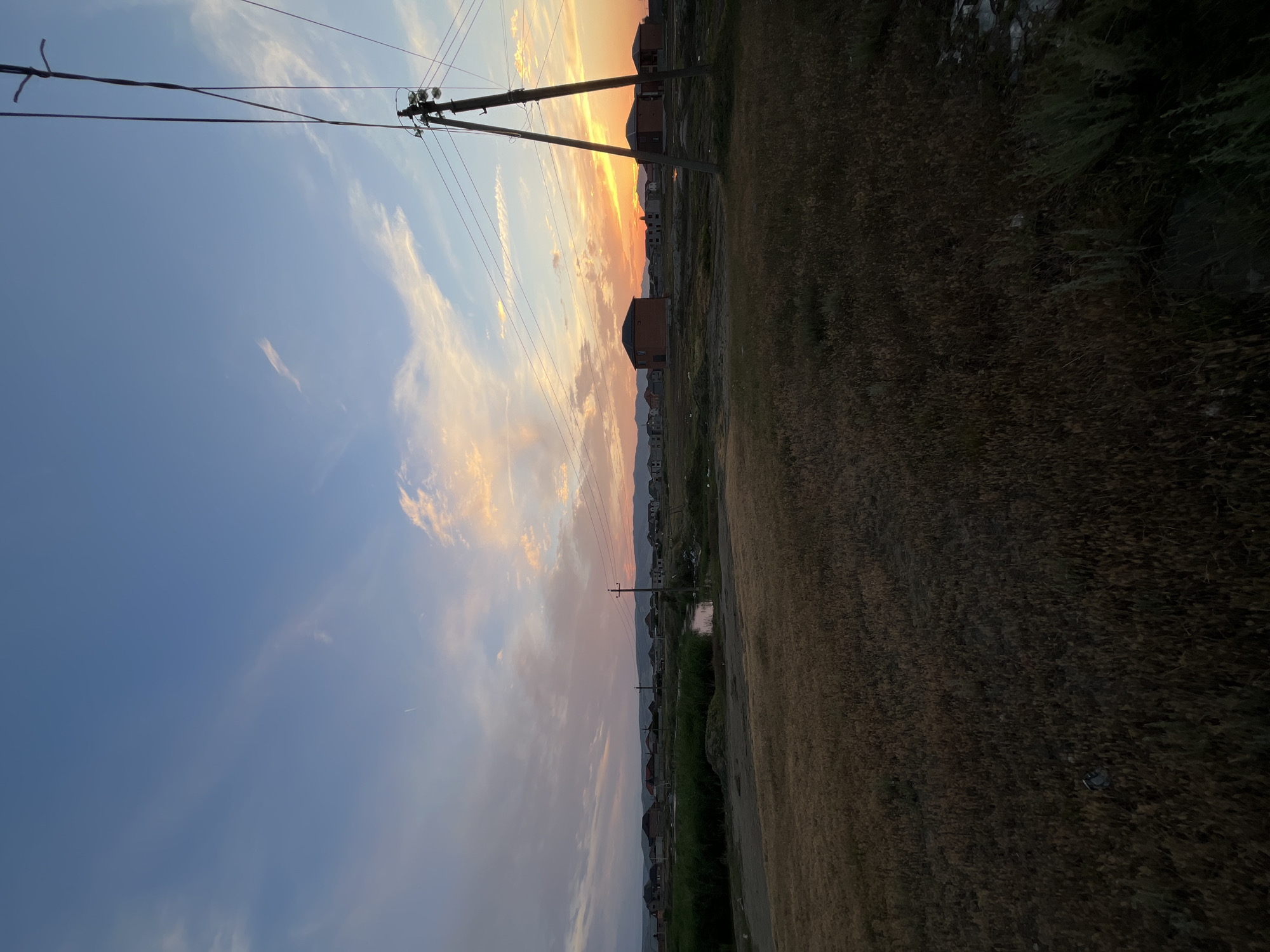 The image depicts a rural landscape at sunset, possibly in the vicinity of Kaspiysk, Russia, based on the provided context. The foreground is dominated by dry, golden grass, suggesting an arid or late-summer environment. In the middle ground, a line of residential buildings is visible, characterized by houses with red-tiled roofs and brick facades. A thin body of water, possibly a canal or stream, runs through the settlement. Power lines and a utility pole are prominent in the right half of the frame, indicating infrastructure in the area. The sky above is a canvas of twilight colors, with hues of blue, pink, and orange, illuminated by the setting sun. Wispy clouds streak across the sky, catching the last rays of light. The overall impression is one of a quiet, developing settlement at the end of the day. There are no people or vehicles visible in the scene.