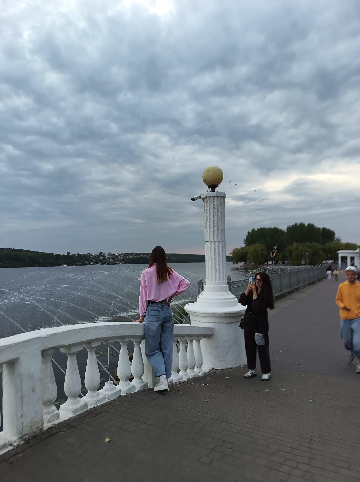 The image depicts a cloudy day scene at the Ternopil Lake, Ukraine. In the foreground, a white balustrade runs along the edge of a promenade. On the promenade, to the left, a young woman with long dark hair, wearing a pink long-sleeved shirt and blue jeans, stands with her back to the camera, looking out at the lake. To her right, another young woman with glasses and dark hair, dressed in a black jacket and dark pants, holds up a smartphone, appearing to take a photo. Further to the right, a person in a bright orange hoodie and blue jeans is seen walking away from the camera, with another figure partially visible behind them. The lake, Ternopil Lake, is a large body of water with a distant shoreline featuring greenery and buildings. White, wavy lines, possibly from water jets or disturbances, are visible on the surface of the lake. A decorative column with a globe-like fixture and a security camera is positioned near the balustrade. The promenade itself is paved with dark rectangular bricks. Trees are visible on the far side of the promenade, and a white structure, possibly a gazebo or shelter, is present in the background.