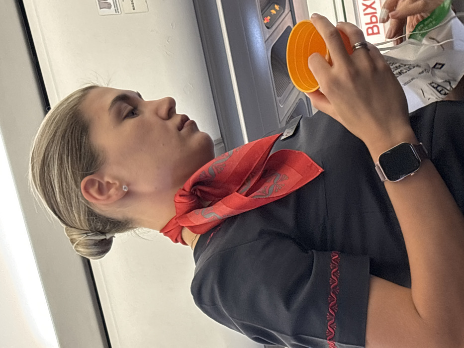 The scene depicts a female individual within an aircraft cabin, contextually located in or operating near Novogorelovo, Russia. The subject is a light-haired female, approximately 20-35 years old, with her hair neatly styled in a bun. She wears a dark, short-sleeved uniform top featuring a red patterned trim on the cuff of her left sleeve, and a red scarf with a repeating grey patterned motif tied around her neck. A dark nametag or badge is clipped to her uniform below the scarf. A small stud earring is visible in her right ear. On her left wrist, she wears a smartwatch with a rose gold-colored rectangular case and a dark mesh band. A simple silver-toned band ring is present on her left ring finger. She is holding an empty or partially filled orange plastic cup in her left hand, while her partially visible right hand appears to be manipulating a white plastic bag or wrapper. Her gaze is directed downwards and to her right. The background consists of the typical white overhead panels and storage compartments of an airplane interior. An overhead passenger service unit with air vents, call buttons, and an illuminated "no smoking" indicator is visible. A white sign with red Cyrillic text "ВЫХОД" (Exit) is visible on a bulkhead or door frame to the right.