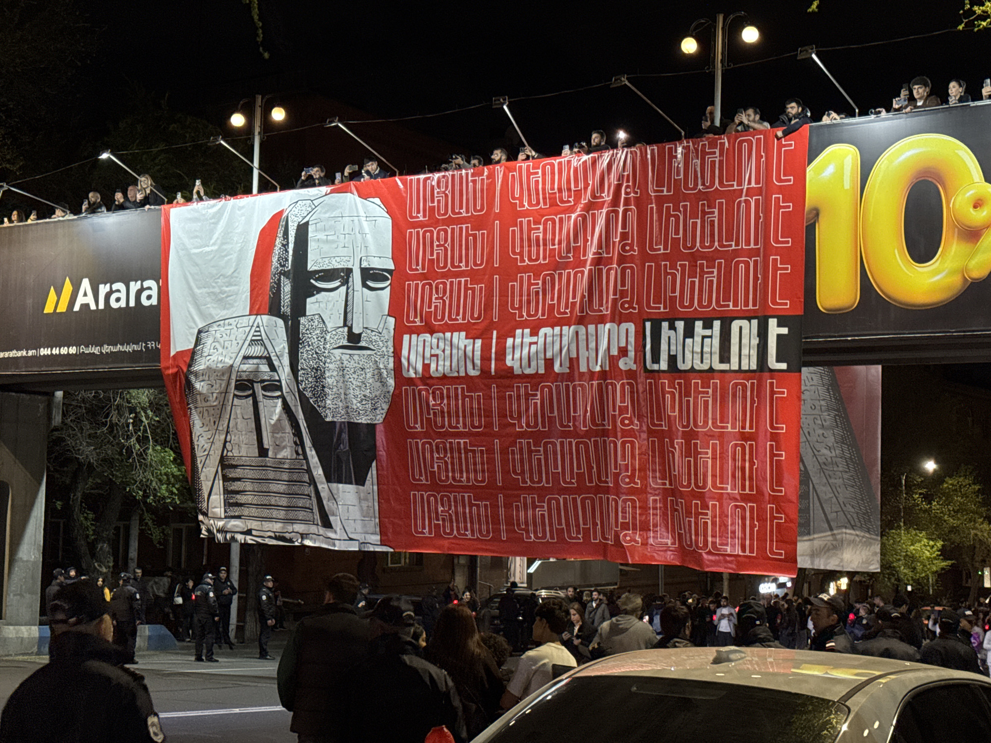 The image depicts a nighttime scene in Yerevan, Armenia, focused on a large banner prominently displayed above a street. The banner, predominantly red with a white vertical stripe on the left, features a black and white illustration of the "We Are Our Mountains" monument, a symbolic representation of Artsakh (Nagorno-Karabakh). Repeated across the red section of the banner in white Armenian script is the phrase "ԱՐՑԱԽ | ՎԵՐԱԴԱՐՁ ԼԻՆԵԼՈՒ Է", which translates to "Artsakh | Return will happen."
A crowd of people has gathered below the banner, with several individuals and police officers visible in the foreground and background. Many people on an elevated structure (likely a pedestrian bridge or part of a billboard frame) are also observing the scene, holding up phones, possibly recording.
To the left of the banner, a large billboard for "Araratbank" is visible, showing the "Ararat" logo, the website "araratbank.am", a phone number "044 44 60 60", and the Armenian text "Բանկը վերահսկվում է ՀՀ Կ" (Bank is supervised by CB of RA). A large yellow "10" graphic is partially visible on the right side of the billboard. The scene is illuminated by streetlights and the bright billboard, confirming it is nighttime. A white car is partially visible in the lower foreground. The presence of the crowd, police, and the banner with its clear message suggests a public gathering or demonstration related to the Artsakh issue.