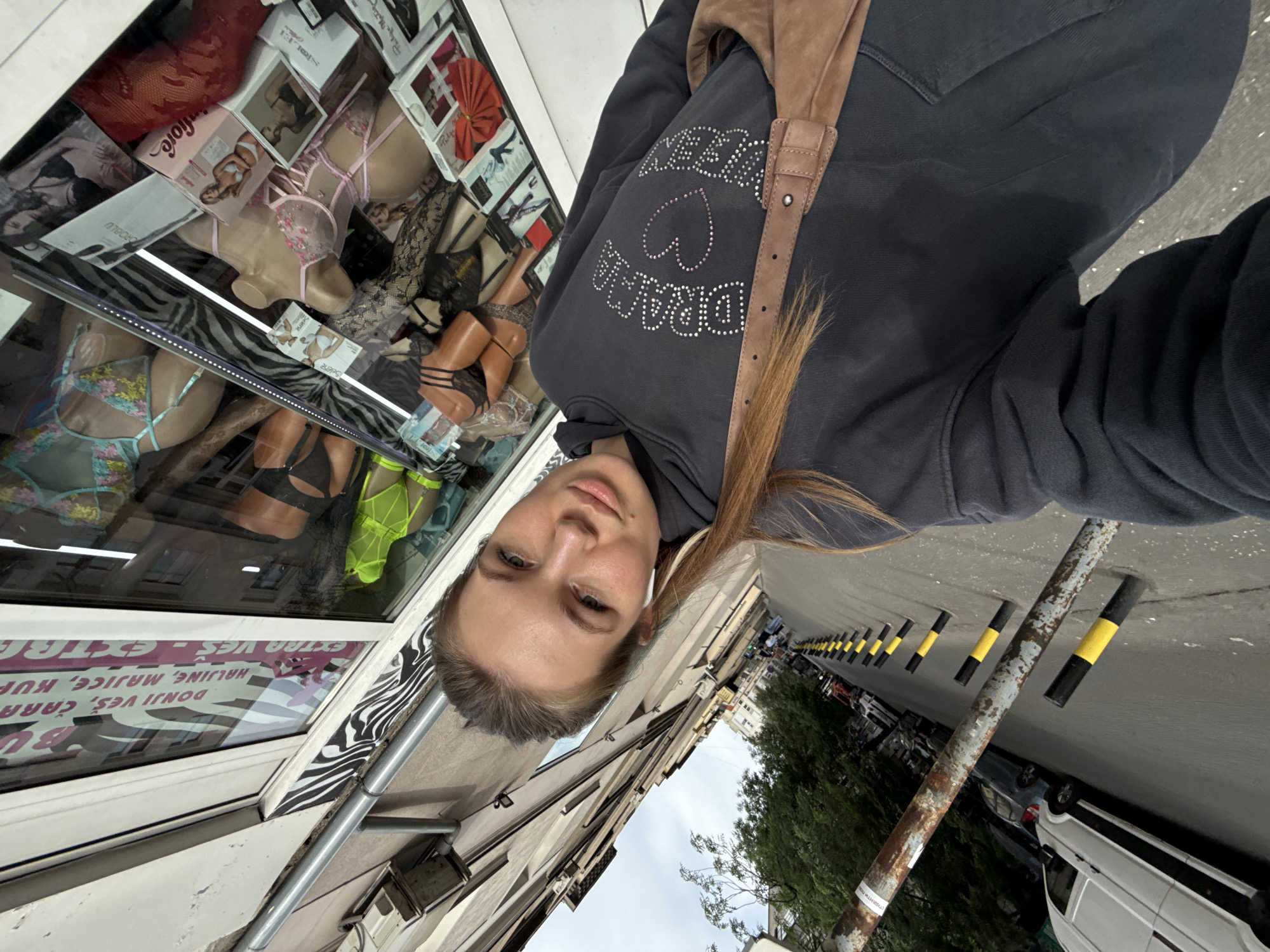 A selfie captured from a slightly tilted angle features a fair-skinned young woman with blonde hair, wearing a dark grey hooded sweatshirt adorned with rhinestone lettering and a heart design. She has a brown leather bag slung over her shoulder and is wearing wireless earbuds. The backdrop is a shop window displaying lingerie mannequins and packaging. The window's signage, in a language other than English, partially obscured by reflections, mentions various clothing items and brands.
The scene appears to be a street in the Vracar Urban Municipality, Serbia. Visible on the street are bollards with black and yellow stripes, parked cars, a white van, and trees. The sky appears cloudy. The photo seems to have been taken on a sidewalk or pedestrian area adjacent to the street.