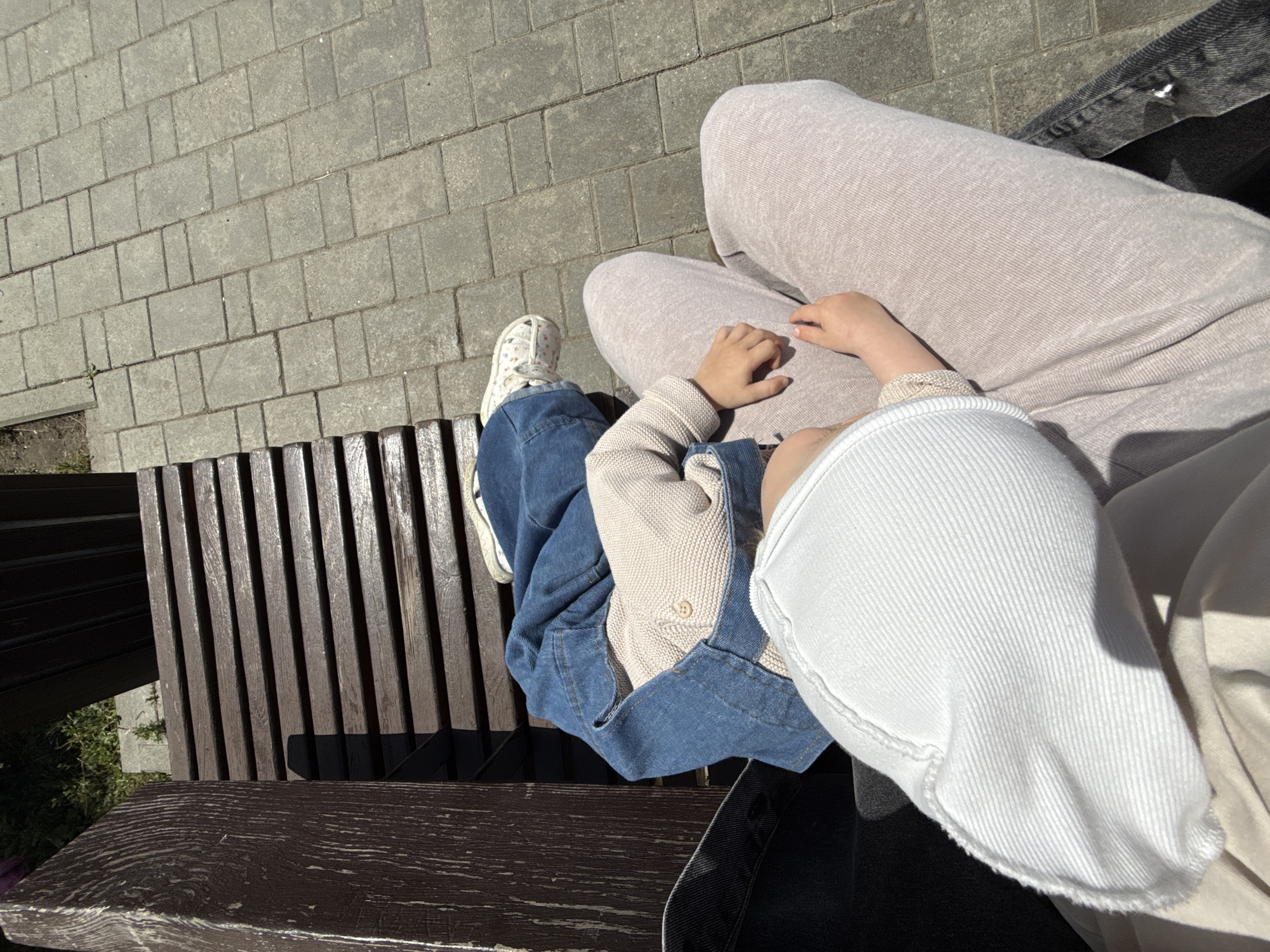 The image shows a top-down view of two people sitting on a park bench. One person, likely an adult, is wearing light beige sweatpants and a light beige top. The other person, a child, is wearing a denim jacket over a beige top and blue jeans. The child's right hand is resting on the adult's lap. The bench is made of dark wood with vertical slats. Behind the bench, a paved walkway made of grey rectangular bricks is visible, with some patches of grass and dirt on the side. The lighting suggests it is daytime, with the sun casting shadows. The scene appears peaceful and relaxed. There is no visible text in the image. The location context is Vladimir, Russia.