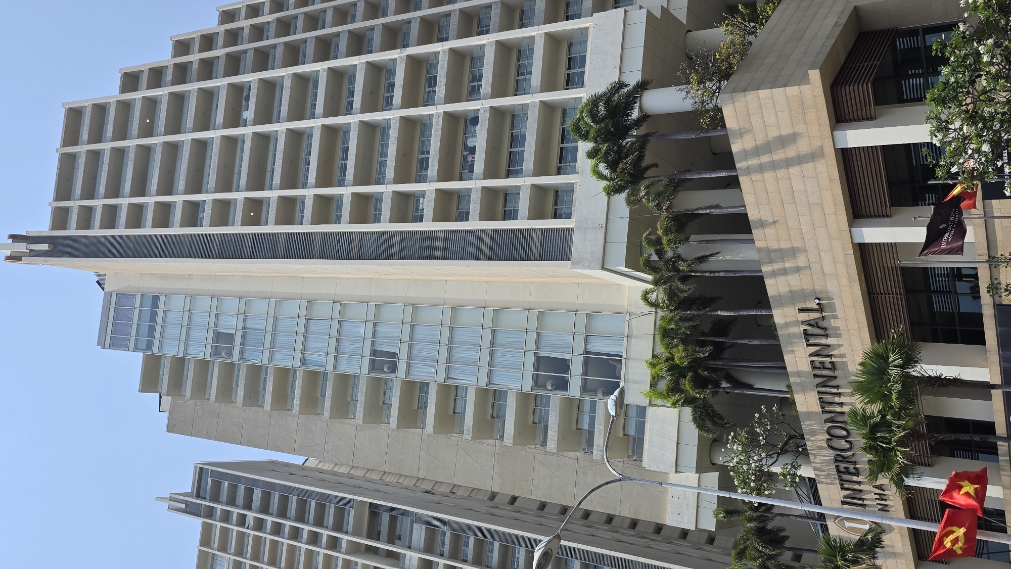 The image shows the InterContinental Nha Trang hotel in Nha Trang, Vietnam. The hotel is a high-rise building with multiple floors and many balconies. The building is a light tan color, and the balconies create a grid-like pattern. In front of the building is a row of palm trees, and a sign displays the hotel's name, "InterContinental Nha Trang". Flags are visible near the hotel's entrance. The sky is clear and blue. The perspective is from a low angle, looking up at the building.