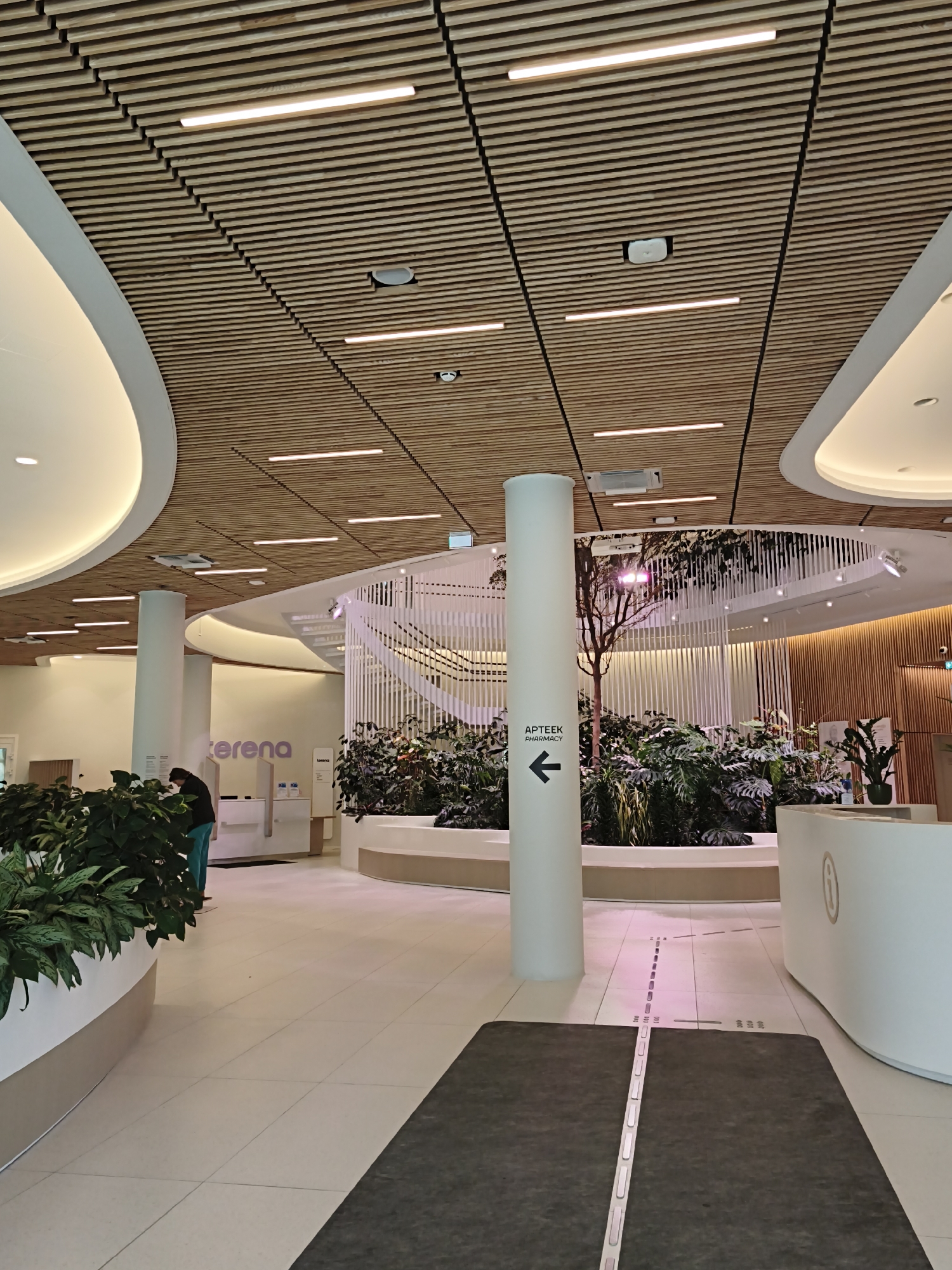 The image depicts the interior of a modern building, likely a public space or a commercial establishment, in Tallinn, Estonia. The architecture features a distinctive wooden slatted ceiling with integrated linear lighting, creating a warm and contemporary ambiance. In the foreground, a large, white cylindrical pillar stands, bearing a black sign with white text that reads "APTEEKK PHARMACY" and an arrow pointing to the left.
Further into the space, lush green plants are arranged in planters, contributing to a natural and inviting atmosphere. A white reception desk or counter is visible on the right, with subtle branding. In the background, a person wearing a dark jacket and teal pants is seen interacting with a counter or kiosk. The floor is tiled, with a dark mat in the foreground featuring a luminous strip. The overall impression is one of a clean, well-lit, and thoughtfully designed interior, possibly a pharmacy, clinic, or a modern retail space. The building's design suggests a focus on natural materials and ambient lighting, typical of contemporary Scandinavian or Baltic architectural trends.