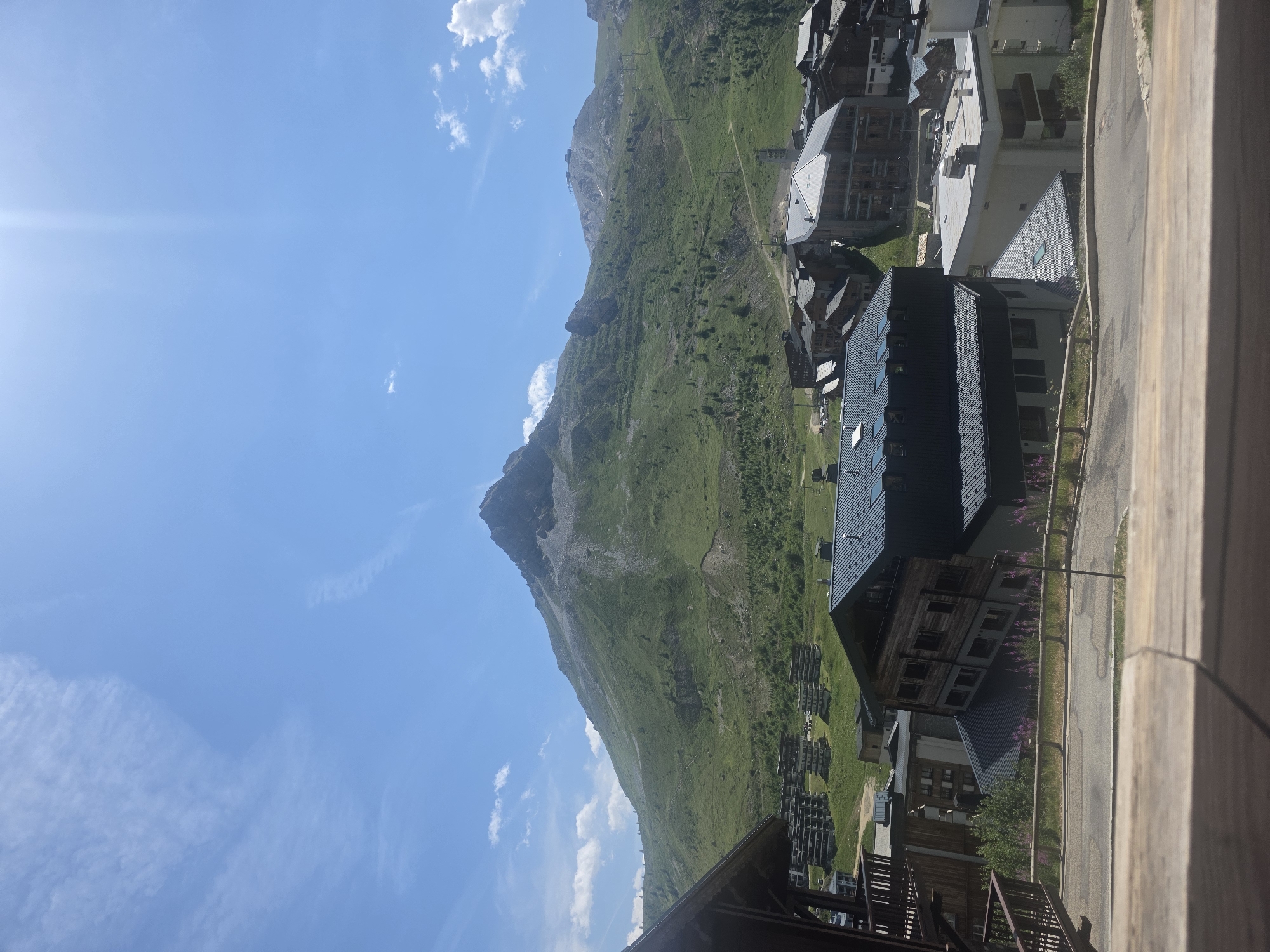 The image depicts a mountainous landscape in Tignes, France, on a clear, sunny day. In the foreground, a wooden railing suggests a balcony or deck, with a blurred wooden surface in the immediate foreground. Below this, a road curves through a village characterized by modern, multi-story buildings with dark, sloped roofs. Some buildings are made of wood, while others have white facades. Purple flowers grow along a wooden fence bordering the road.
In the middle ground, more buildings of varying architectural styles are visible, nestled on the lower slopes of a large mountain. These buildings appear to be part of a ski resort, with some featuring multiple balconies. The mountain itself dominates the background, its green slopes dotted with scattered trees and evidence of ski lifts or trails. The peak of the mountain is rocky and rugged. The sky is a bright blue with a few wispy white clouds and a bright sun flare in the upper right quadrant. The overall scene conveys a sense of a summer day in a mountainous alpine resort.