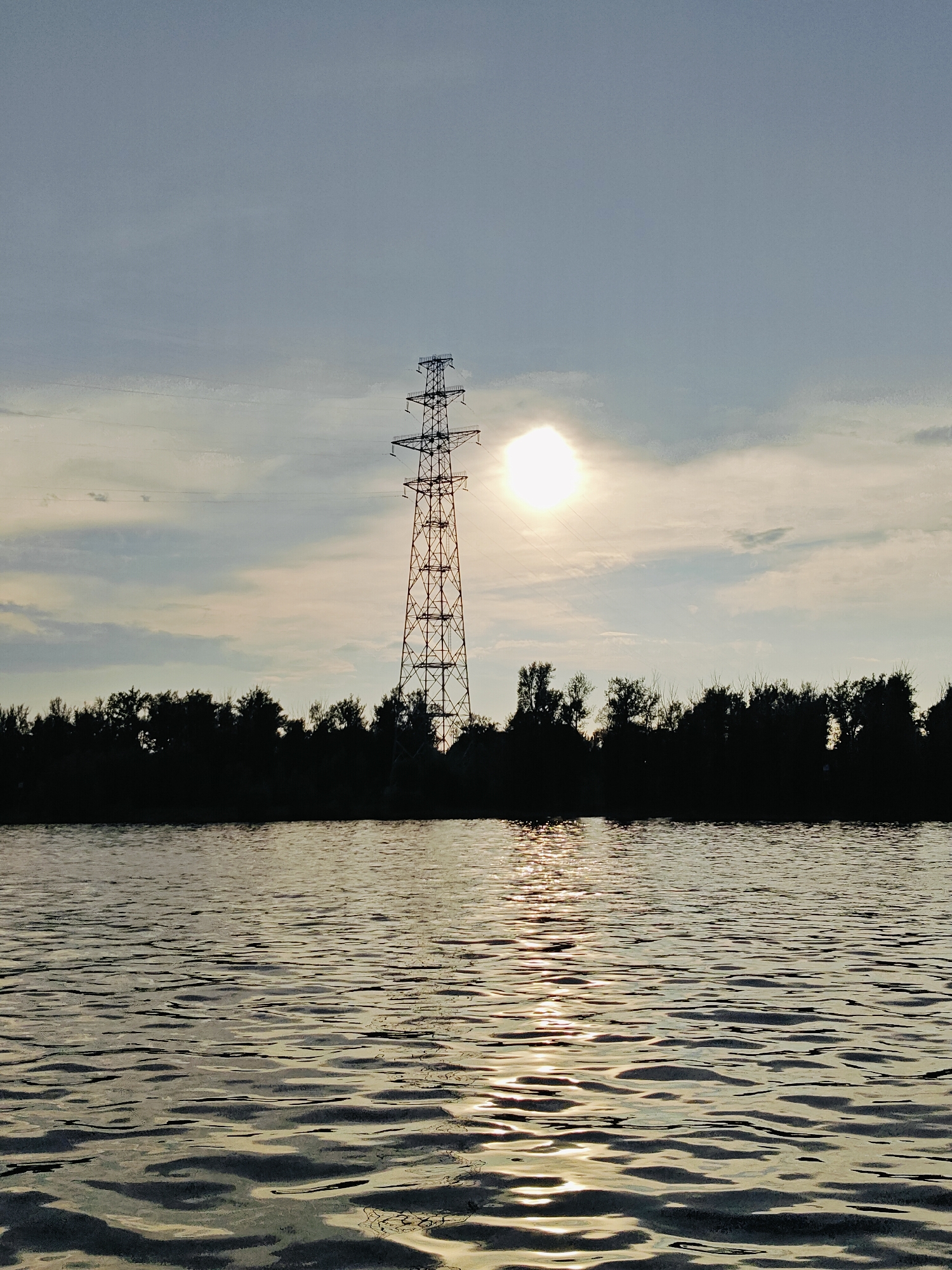 This photograph depicts a silhouetted electrical pylon situated near the city of Noviy, Russia. The pylon stands tall in the distance, positioned behind a dark line of trees that form a dense forest. The sun, partially obscured by clouds, is visible in the upper right portion of the sky, casting a bright glare. In the foreground, a body of water reflects the sunlight and the silhouettes of the trees and pylon, creating shimmering patterns on its surface. The overall scene is serene, with a muted color palette dominated by grays, blues, and blacks, indicative of either dawn or dusk. There are no people or animals visible in the image.