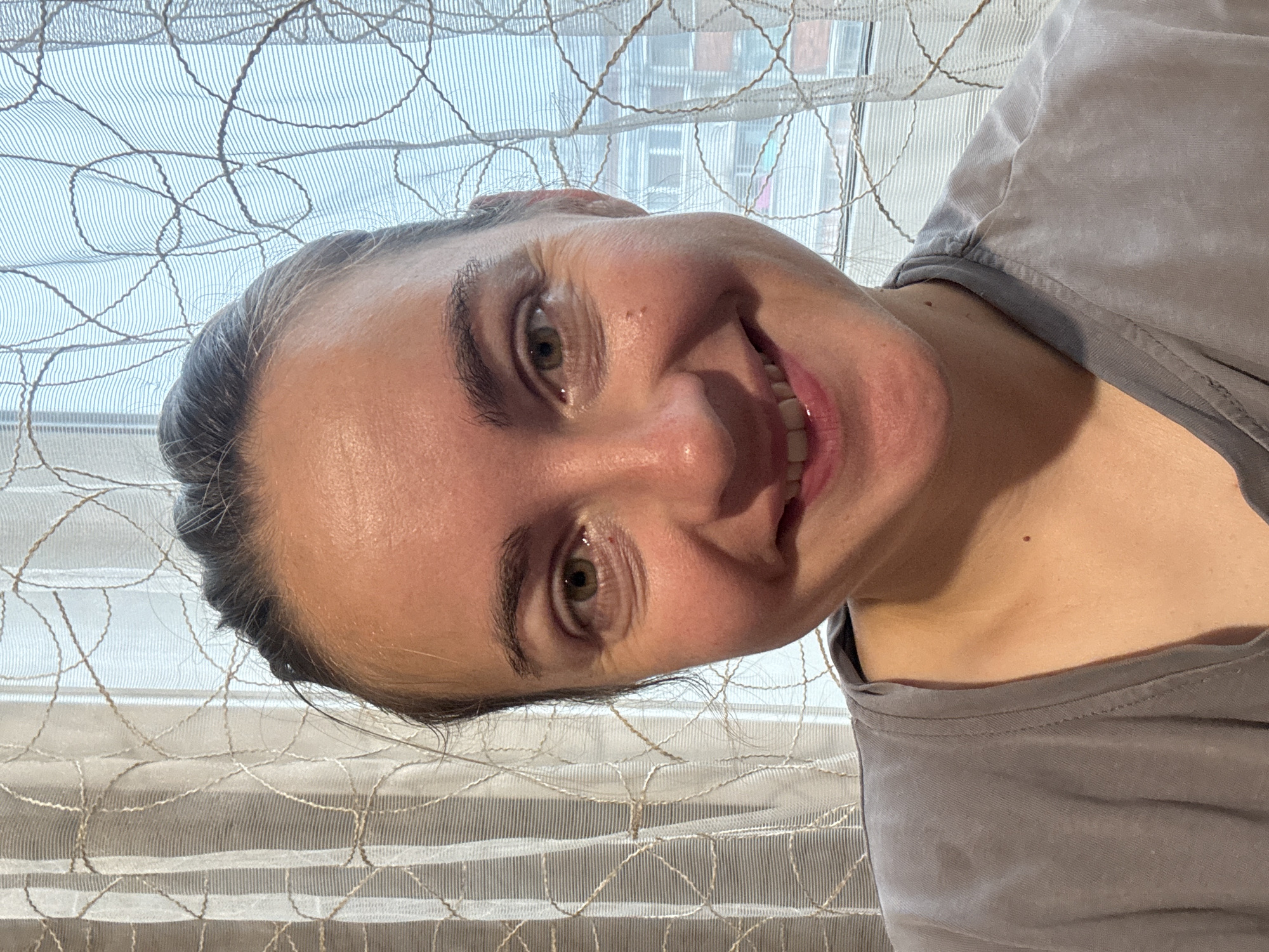 A medium shot, selfie-style portrait of a young adult woman with dark hair pulled back, smiles into the camera. She is wearing a grey t-shirt. The background consists of a sheer, white, patterned curtain and a window with a view of buildings.
