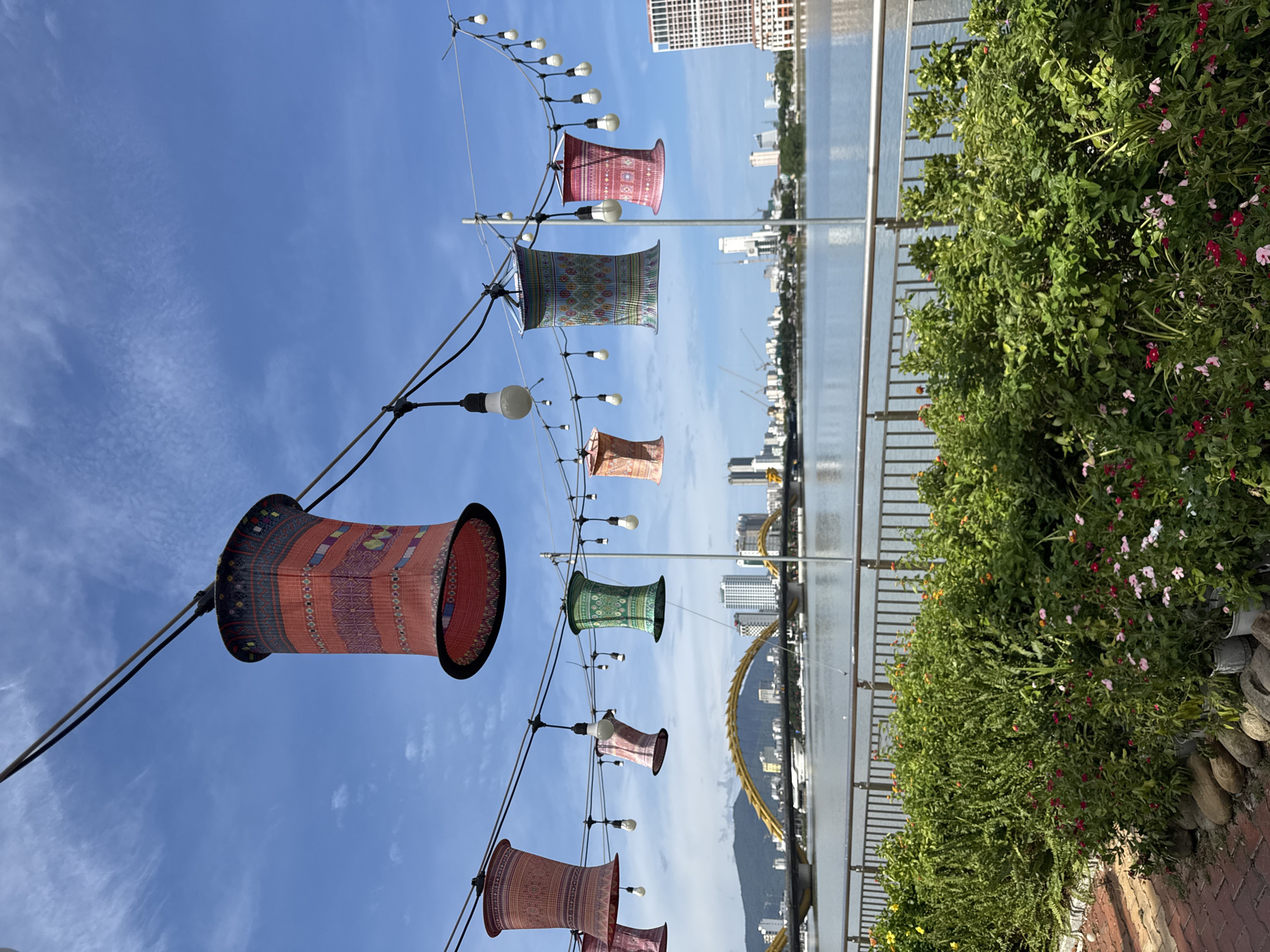 The image captures a scenic view of the Han River in Đà Nẵng, Vietnam, with the iconic Dragon Bridge prominently featured in the background. The foreground is adorned with lush green bushes and vibrant flowers, creating a picturesque setting. Overhead, a string of decorative lanterns and light bulbs hangs, adding a festive ambiance to the scene. The sky above is a brilliant blue, dotted with scattered white clouds, further enhancing the overall beauty of the landscape. The city skyline, with its modern buildings and cranes, forms a striking contrast with the natural elements in the foreground.