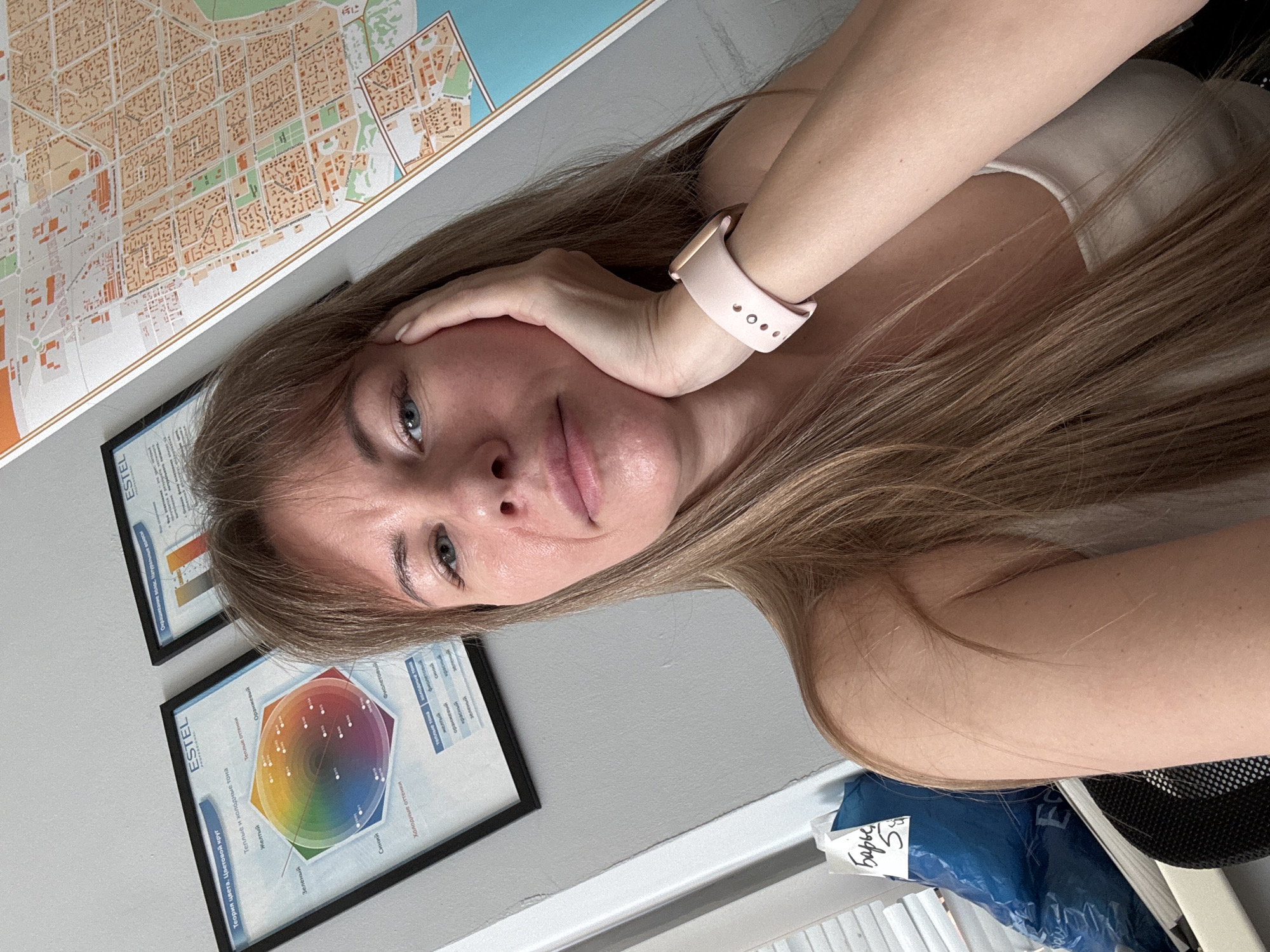 A woman with blonde hair is sitting at a desk. She is wearing a light-colored tank top and a pink watch. Her right hand is touching her face. Behind her are two framed posters, one of which has the word "ESTEL" on it. The second poster seems to describe color theory. A large map is also visible above the posters.
The text on the map is illegible, so I cannot say if it's a map of Tolyatti, Russia.