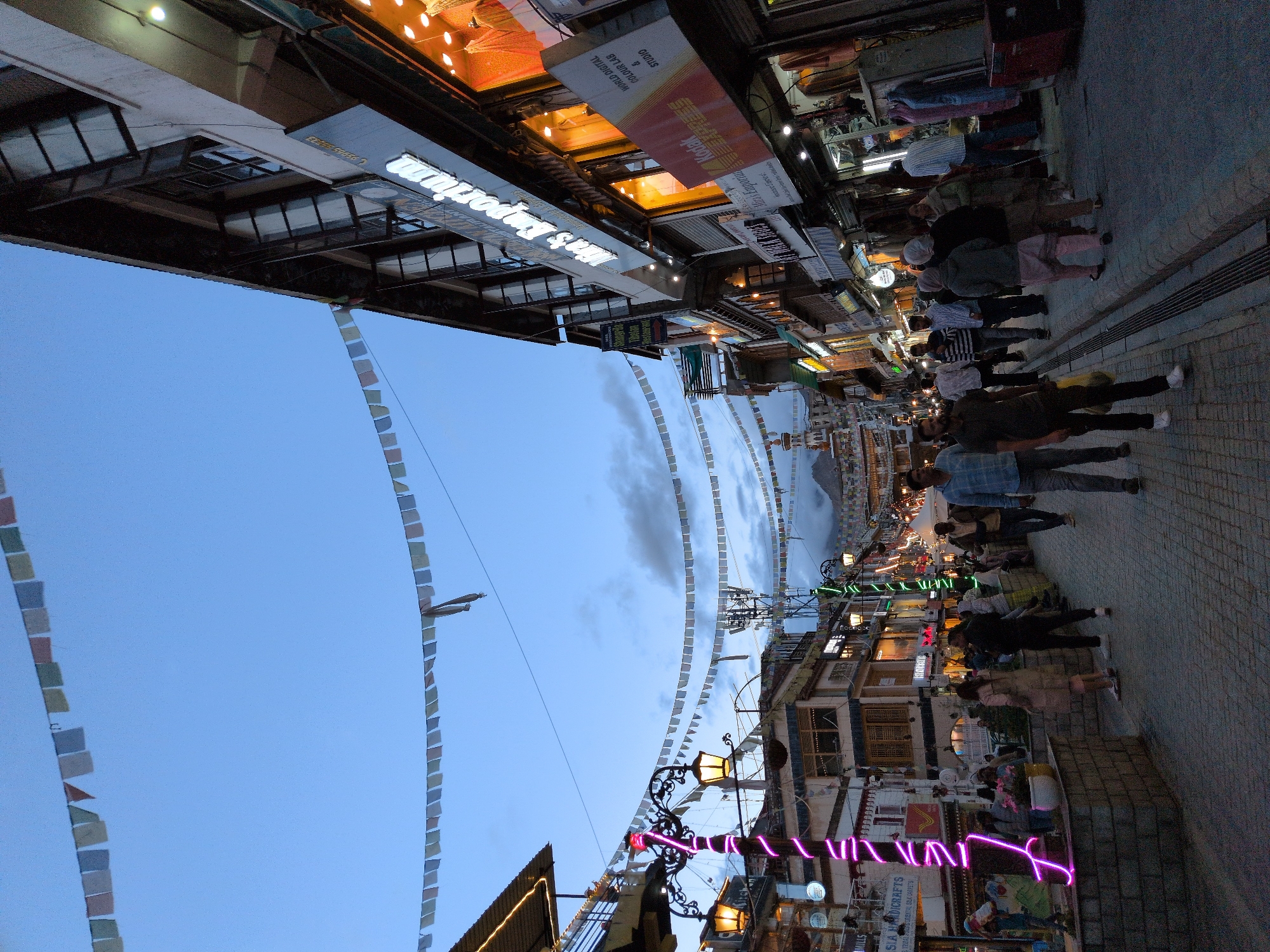 This is a ground-level, wide-angle, vertical shot of a street in Leh, India, likely taken in the early evening. The sky is a deep blue, with some clouds visible. Prayer flags are strung across the street overhead, some appearing faded. The street is paved with cobblestones and lined with multi-story buildings that house shops. The buildings have a traditional architectural style, with wooden balconies and intricately carved details visible.
The shops are illuminated by warm interior lights and some neon signage, giving the scene a vibrant yet serene atmosphere. One prominent sign reads "Men's Emporium." Other signs suggest businesses like "Asia Handicrafts," "Tibetan Restaurant," and "Kodak Express Colour Lab & Studio."
There are several people walking along the street, some individually and some in small groups. They are dressed in casual clothing suitable for the climate. One person is carrying a yellow bag. A decorative street lamp, wrapped in pink neon lights, stands prominently on the left side of the street. The overall impression is of a bustling but orderly marketplace, typical of Leh's main thoroughfares.