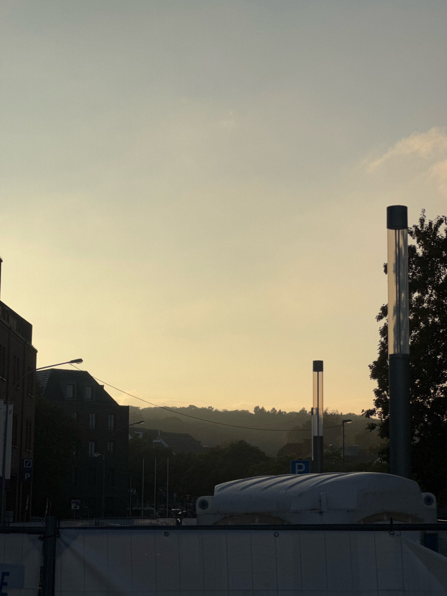 The image depicts a street scene in the city of Cleves, Germany, during what appears to be sunset or early morning. The sky is a pale yellow-orange hue, suggesting a low sun angle. In the foreground, a white, curved structure, possibly a portable toilet or a part of construction, is visible, with a metal barrier in front of it. To the right, a tall, cylindrical object with a dark top and a reflective metallic surface stands prominently, with a tree silhouette behind it. To the left of this object, another, shorter similar cylindrical structure is visible. Behind these elements, several dark buildings are silhouetted against the sky, including a building with a pitched roof and multiple windows, and a taller brick building on the far left. Streetlights are present, one illuminating the area near the pitched-roof building. Further in the background, a hazy, tree-covered hillside slopes upwards, with some indistinct buildings visible on its slopes. A blue parking sign with a white "P" is discernable near the middle ground. The scene conveys a sense of urban twilight with a slightly industrial or construction feel due to the foreground elements.