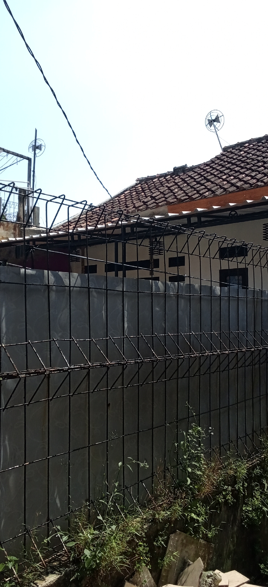 The image captures an outdoor scene in Banjaran, Indonesia, focusing on a black metal fence in the foreground. Beyond the fence, a building with a tiled roof is visible. The roof tiles are reddish-brown and laid in a standard pattern. Above the building, the sky is bright and clear, indicating a sunny day. On the roof, two small satellite dishes are mounted. The fence is made of wire mesh and supports, with some rust visible on the metal. Behind the fence, a grey, textured wall forms the facade of the building. Some green vegetation, including grass and small plants, is growing along the base of the fence and wall, suggesting an unkempt area. Electrical wires are strung across the upper left portion of the image, crossing the clear sky. The overall scene depicts a residential or commercial area with typical outdoor elements.