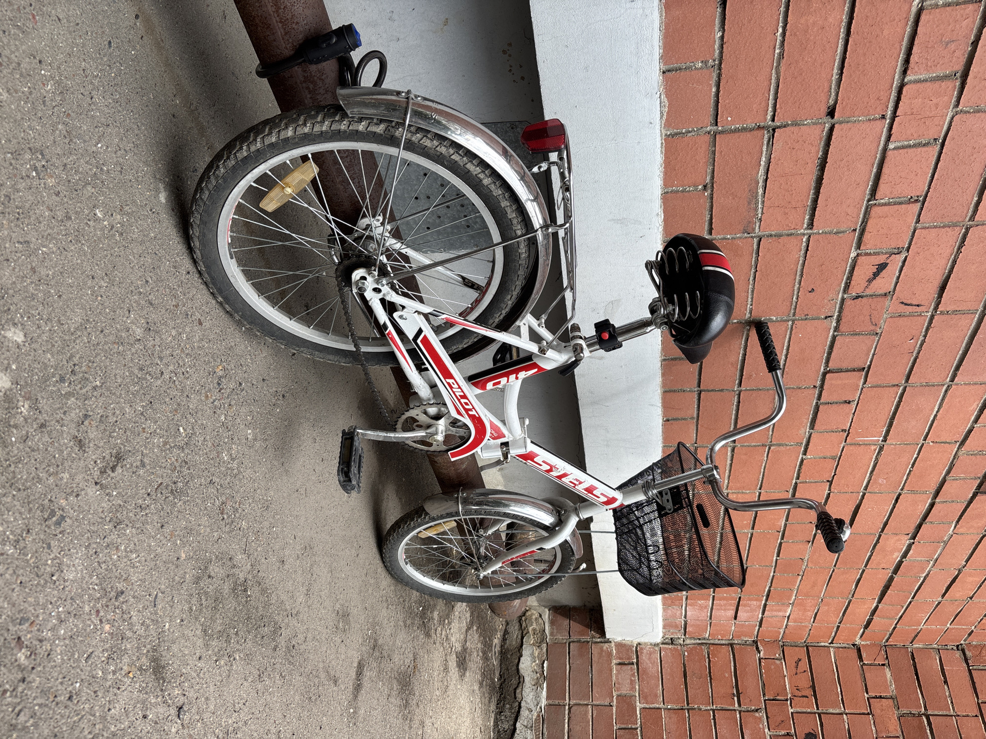 The image shows a white and red Stels Pilot bicycle parked against a red brick wall, next to a white concrete ledge. A large metal pipe runs horizontally in front of the bike, and the bicycle is locked to it with a black U-lock. The bicycle has a black seat with red and white accents, a black wire basket on the handlebars, and a red taillight. The scene appears to be in an urban outdoor setting, likely in the city of Blagoveshchensk, Russia, given the context. There are no people visible in the image, and no actions are taking place. The environment is composed of the brick wall, concrete ledge, metal pipe, and asphalt ground.