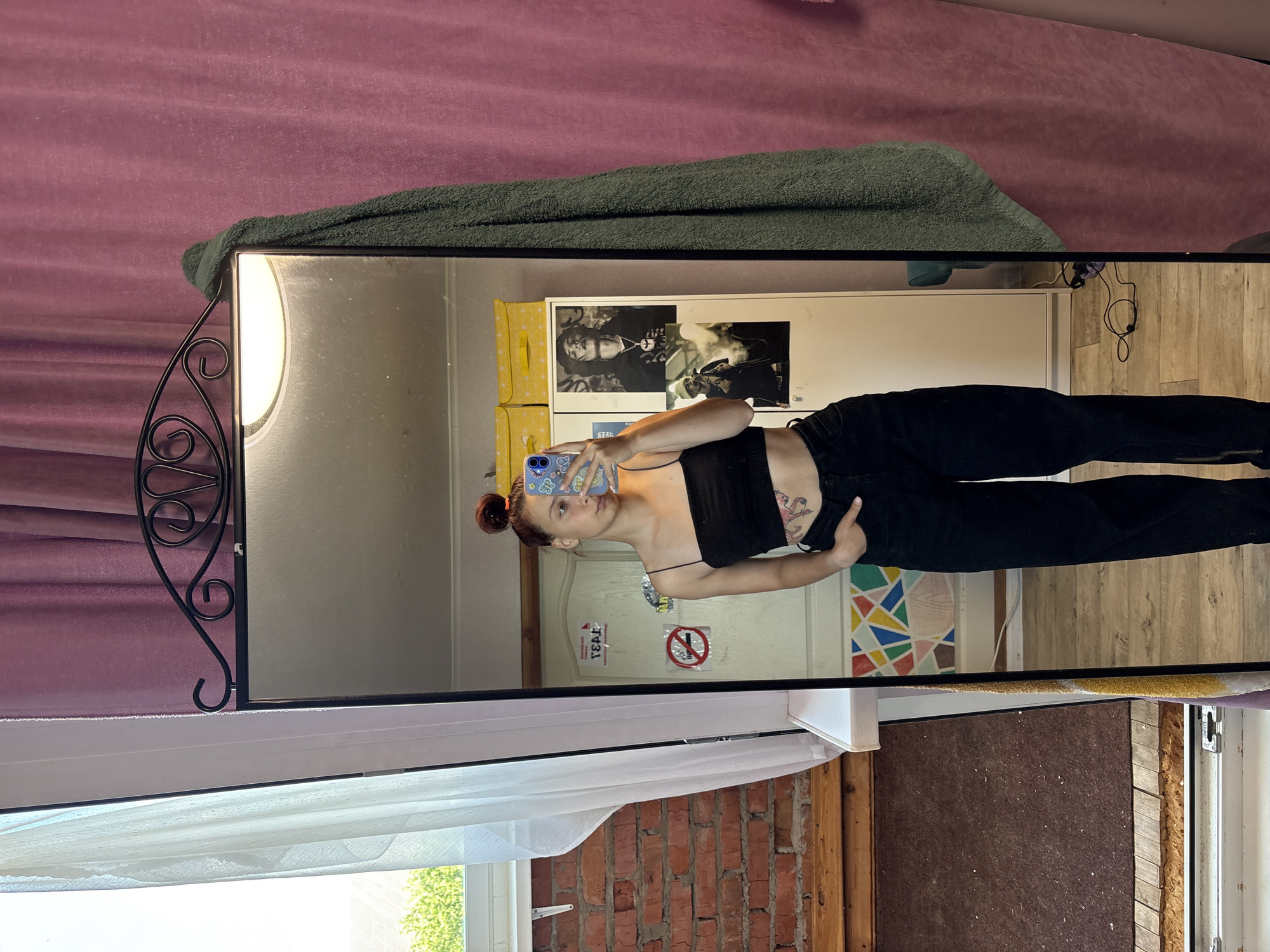 A young woman is captured in a mirror selfie, posing casually in what appears to be a bedroom or living space in Прикамский, Russia. The image is taken from a slightly high angle, reflecting the woman, parts of the room she is in, and elements of the room where the mirror stands.
The primary subject, the woman, stands in front of a large, rectangular mirror with a decorative black wrought-iron style frame on its left side. She has dark hair tied up in a bun and is wearing a black strappy top and black trousers. Several tattoos are visible on her left hip/torso area. She holds a light blue smartphone with a patterned case up to take the photo, looking directly into the phone's camera. Her expression is neutral and relaxed.
In the reflection behind her, a white wall is visible, adorned with posters. One prominent black and white poster features a person with a chain. To the left of this, there are yellow storage boxes. Further back, a door or wall section displays a colorful geometric pattern. On a white surface behind the woman's head, two pieces of text are visible: a red circular "No Smoking" sign (a crossed-out cigarette) and the reversed Cyrillic letters "ТЕАТ", possibly part of "ТЕАТР" (Theatre). The floor in the reflection appears to be light-colored wood.
Outside the mirror's reflection, on the left and top, a textured purple or pink curtain hangs. A dark green towel is draped over the top edge of the mirror frame. Below the mirror, a window with sheer white curtains offers a view of green trees and a building outside, suggesting it is daytime. A section of exposed red brick wall and wooden beams is also visible to the lower right of the mirror. The overall setting suggests an eclectic and lived-in environment.