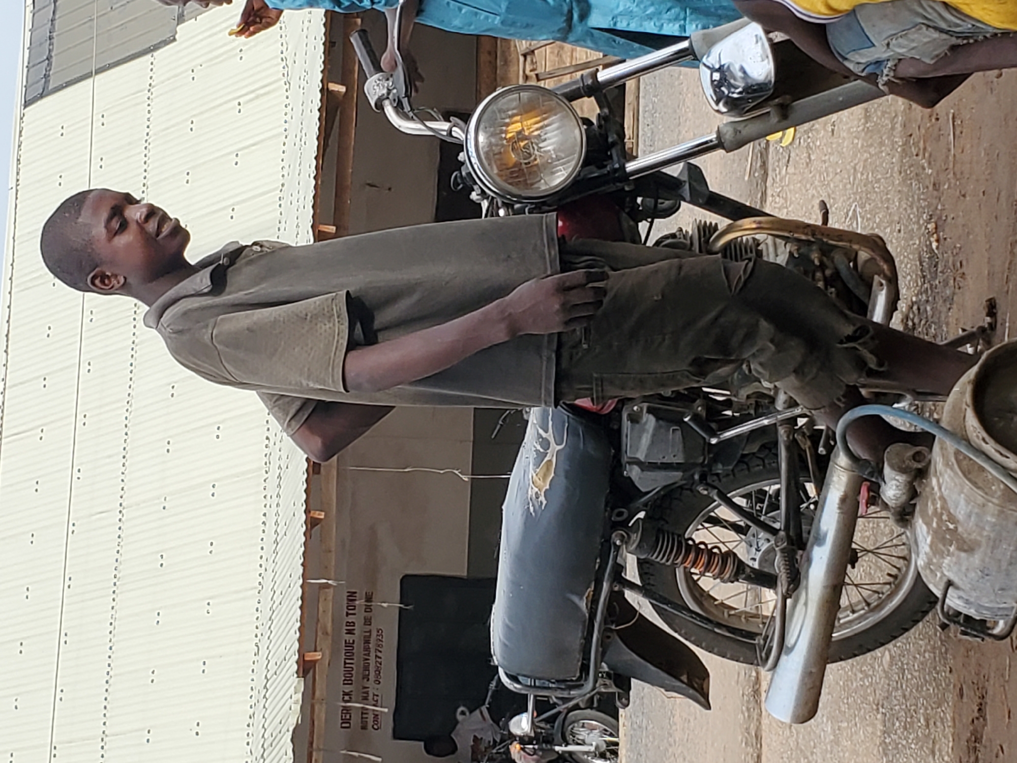 A young man is sitting on a motorcycle in Mararaba, Nigeria, near the "DERICK BOUTIQUE MB TOWN" shop (Contact: 08082778935). The motorcycle is an older model. Two other people are partially visible. The setting appears to be an urban street.